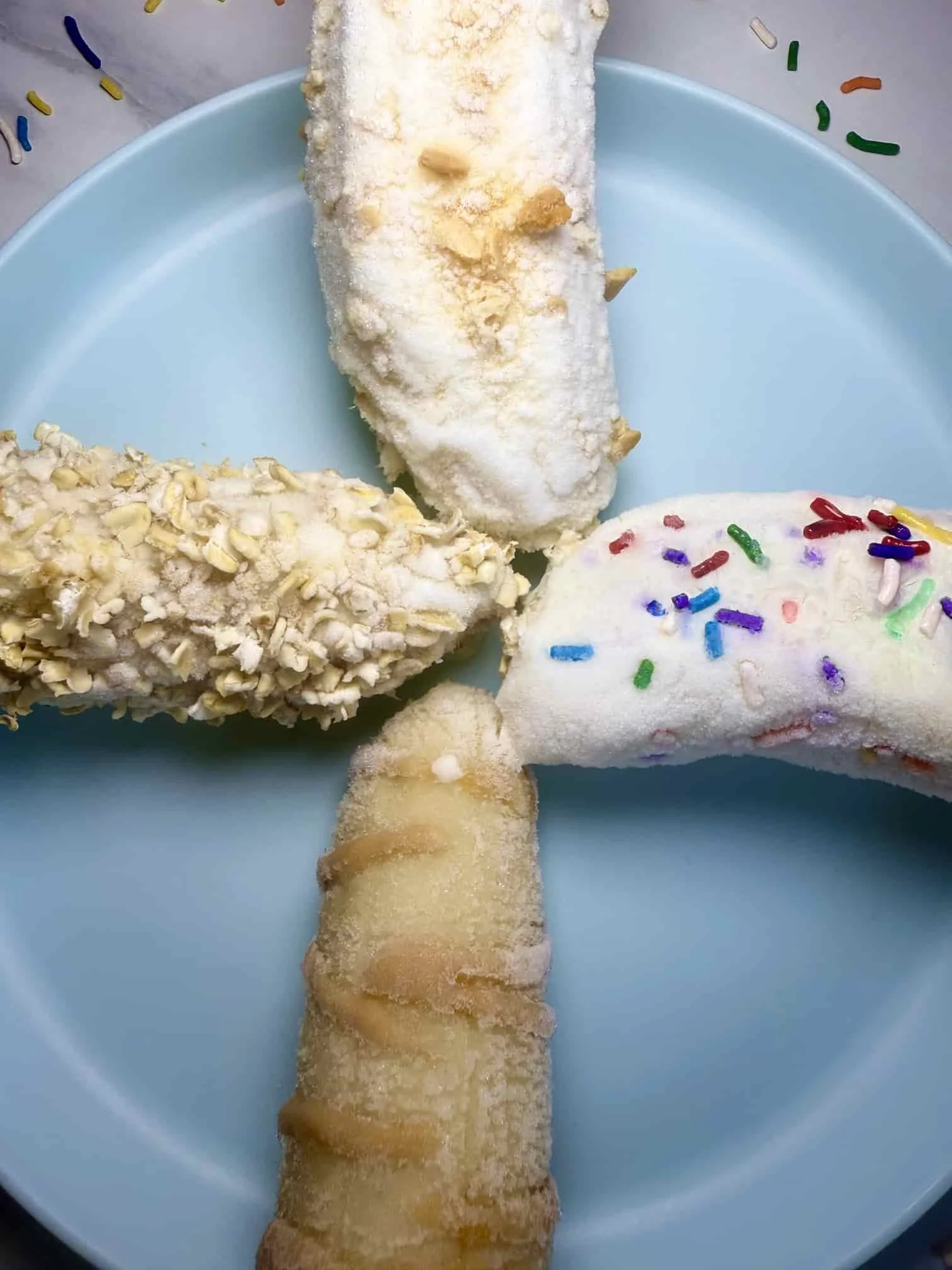 Four banana fruit popsicles with different toppings including sprinkles, oats and peanut butter on serving plate