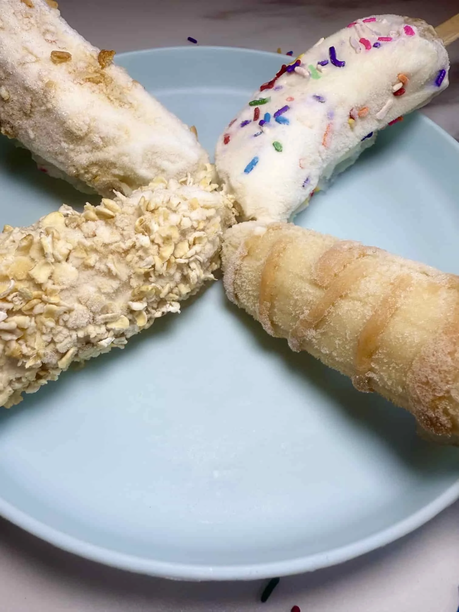 Four banana fruit popsicles with different toppings including sprinkles, oats and peanut butter on serving plate