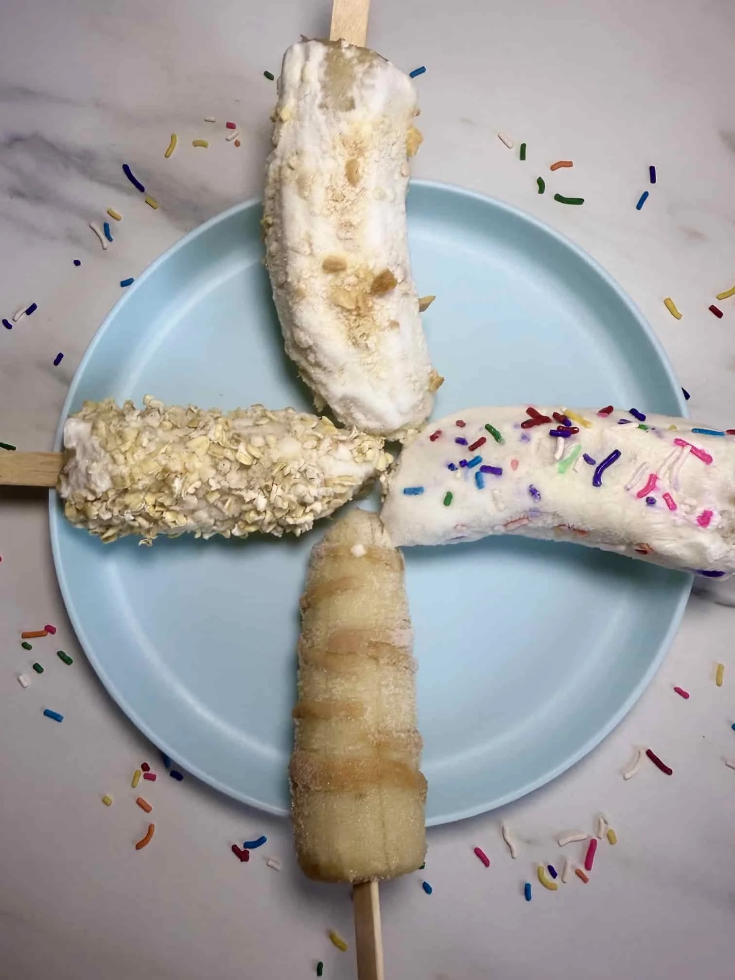 Four banana fruit popsicles with different toppings including sprinkles, oats and peanut butter on serving plate