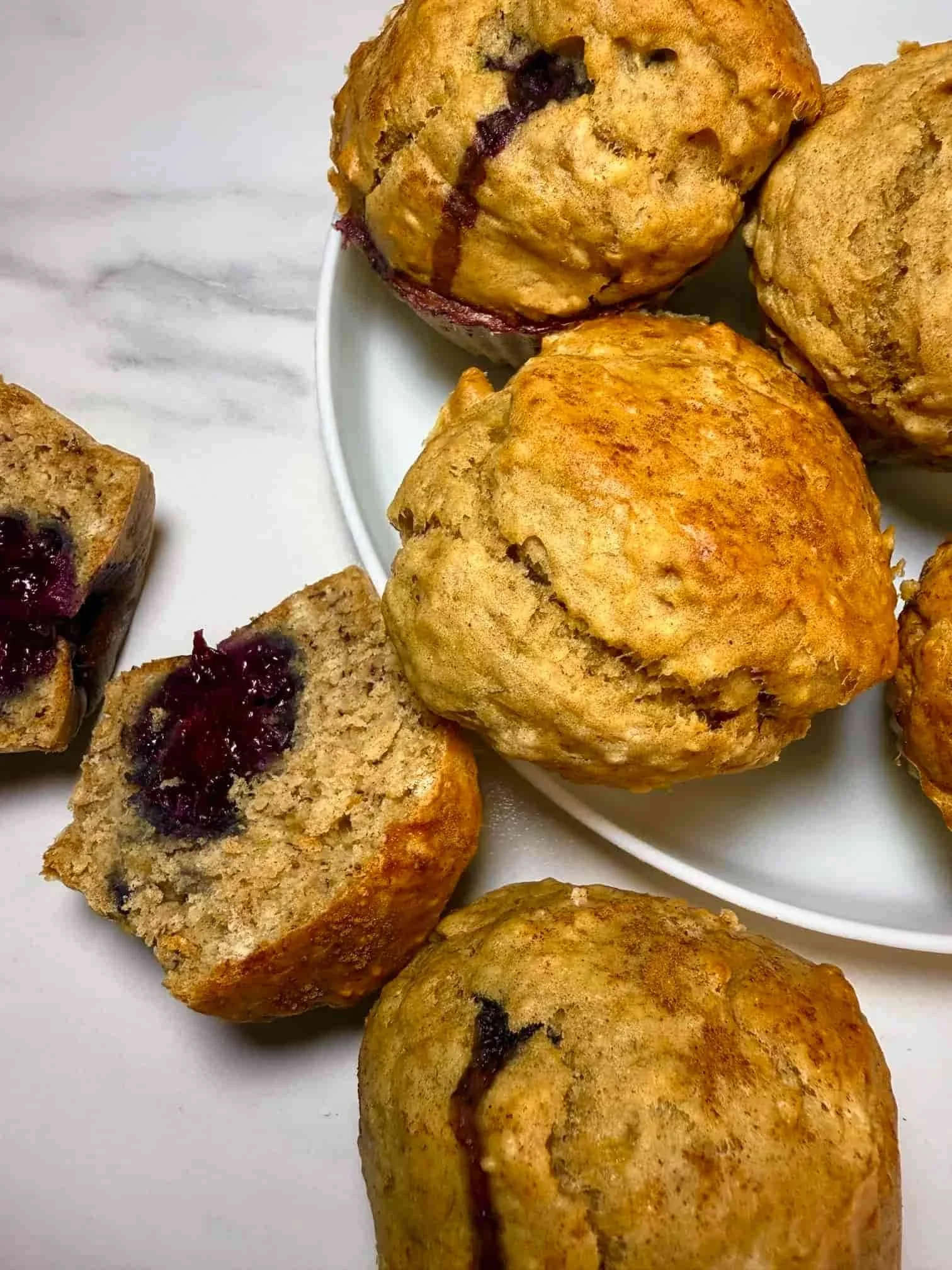 Several baked banana and blackberry muffins on a serving plate, some whole and some cut in half to show the fruit inside