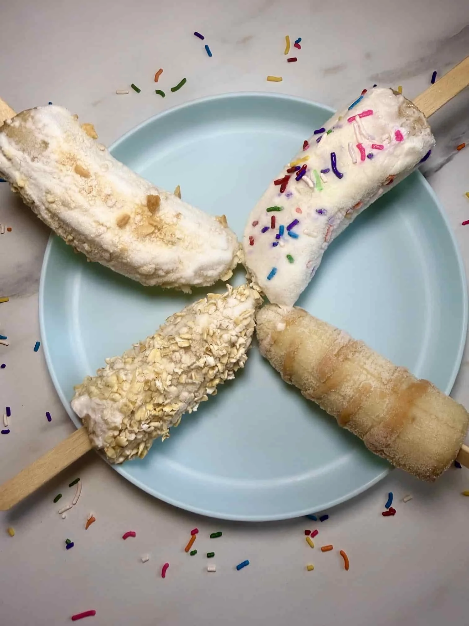 Four banana fruit popsicles with different toppings including sprinkles, oats and peanut butter on serving plate