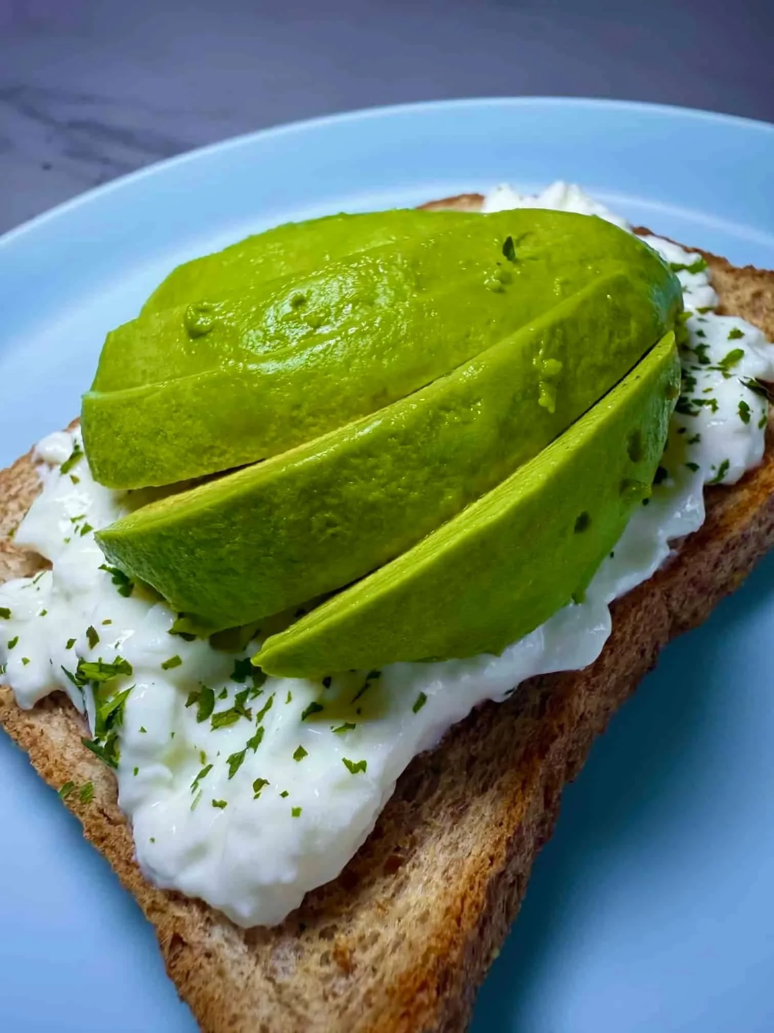 A close up image of slices of avocado on top of toast spread with cottage cheese and herbs