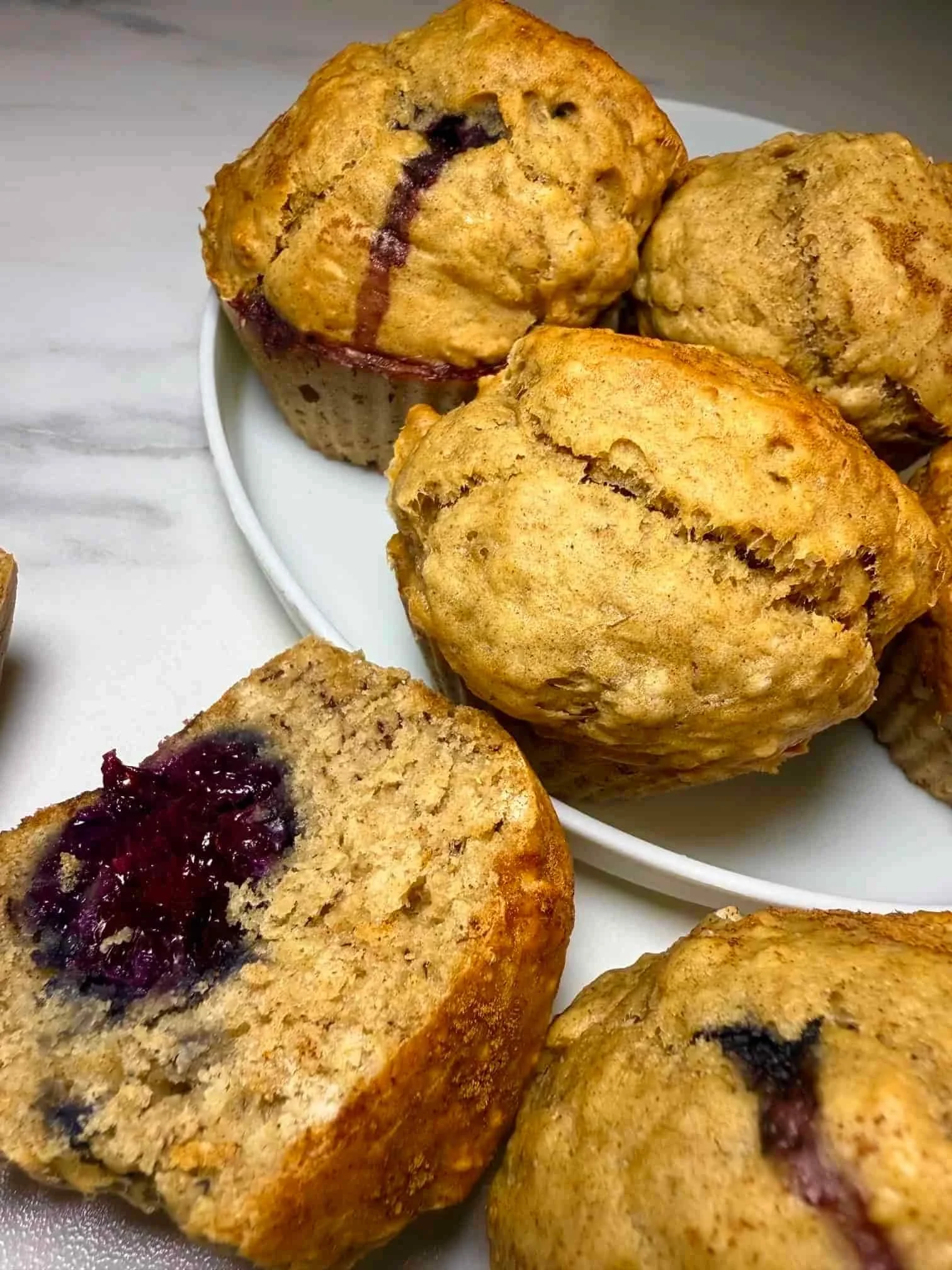 Several baked banana and blackberry muffins on a serving plate, some whole and some cut in half to show the fruit inside