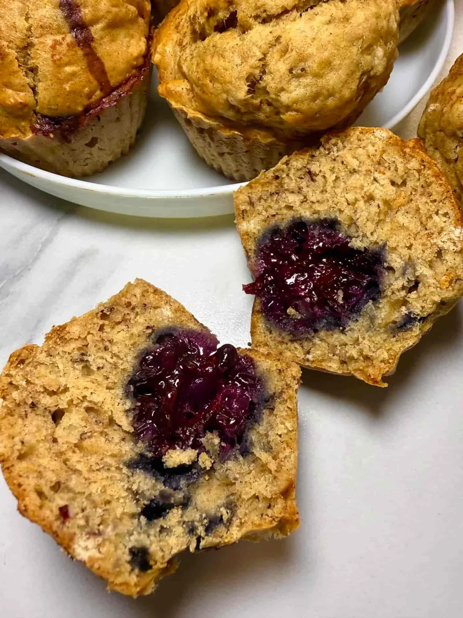 Several baked banana and blackberry muffins on a serving plate, some whole and some cut in half to show the fruit inside