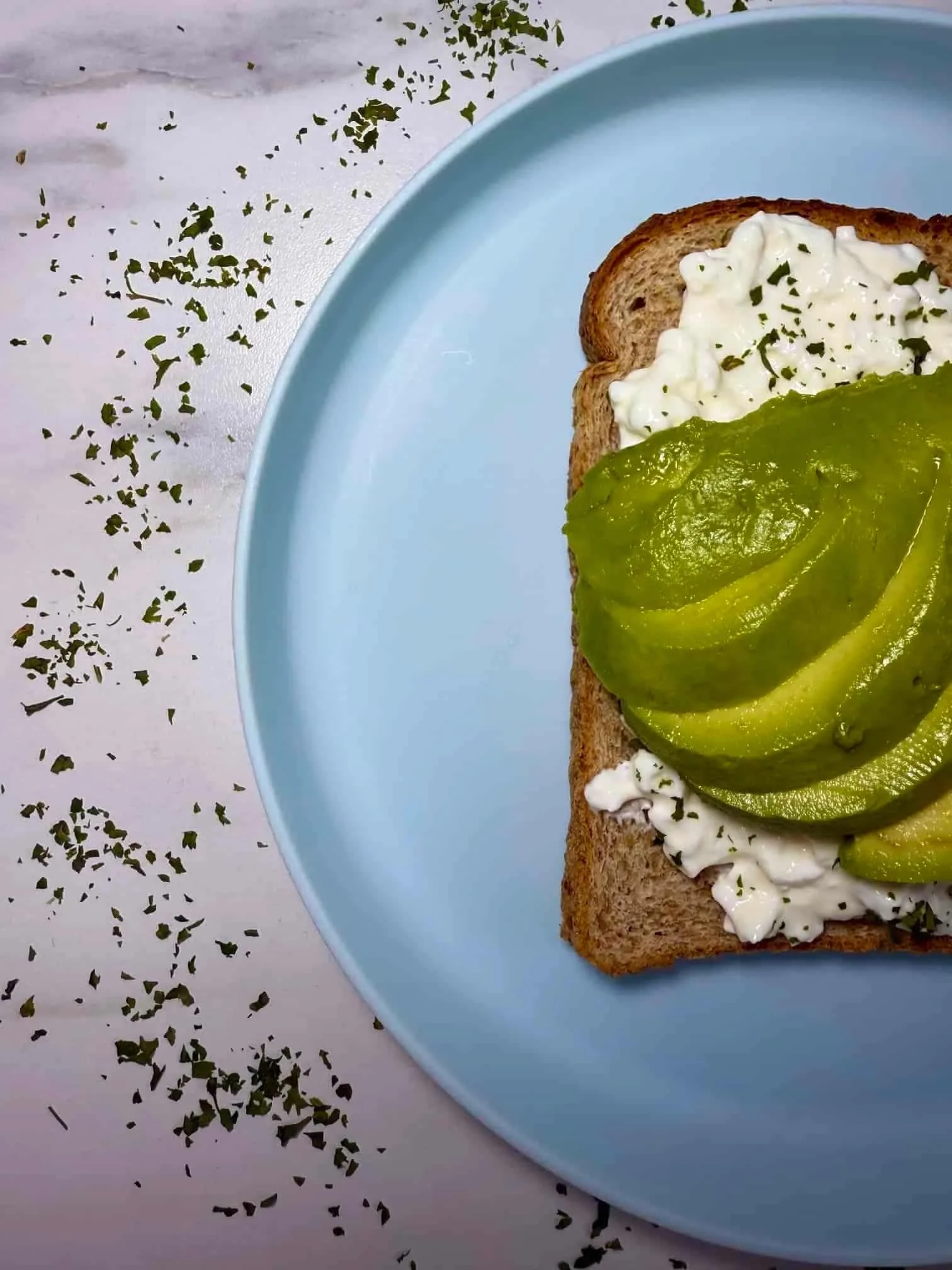 A close up image of slices of avocado on top of toast spread with cottage cheese and herbs