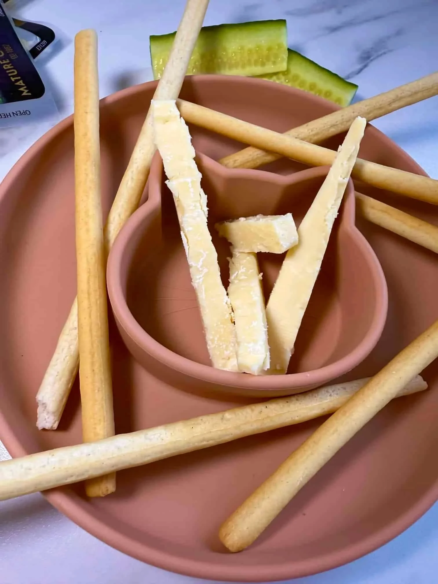 Breadsticks and fingers of cheese together with some cucumber presented on a toddler dish
