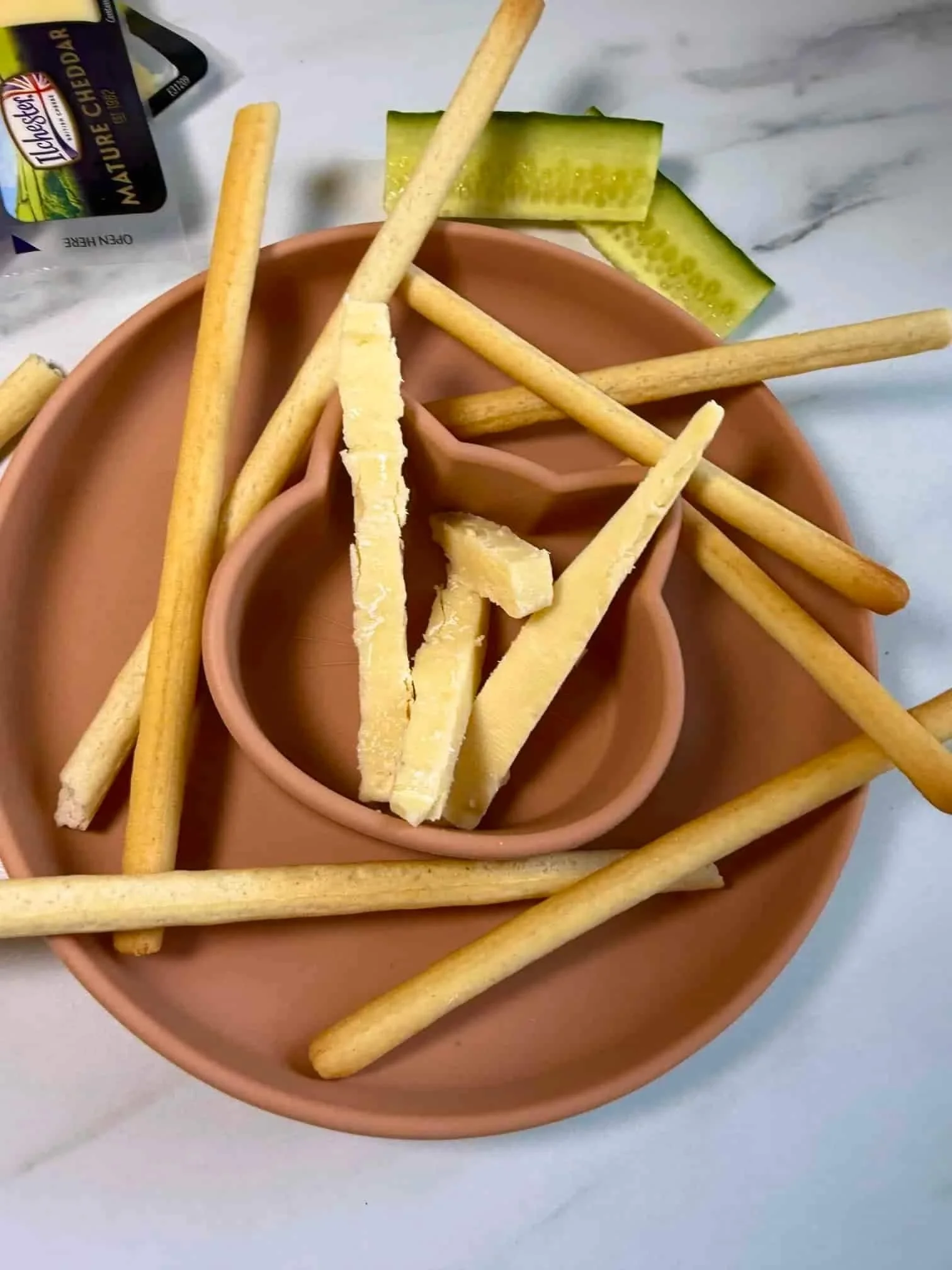 Breadsticks and fingers of cheese together with some cucumber presented on a toddler dish