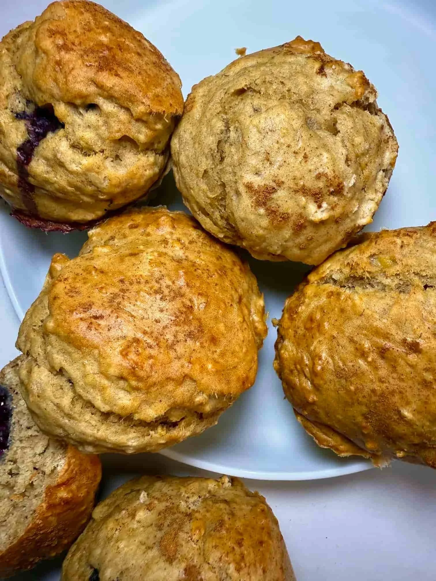Several baked banana and blackberry muffins on a serving plate, some whole and some cut in half to show the fruit inside