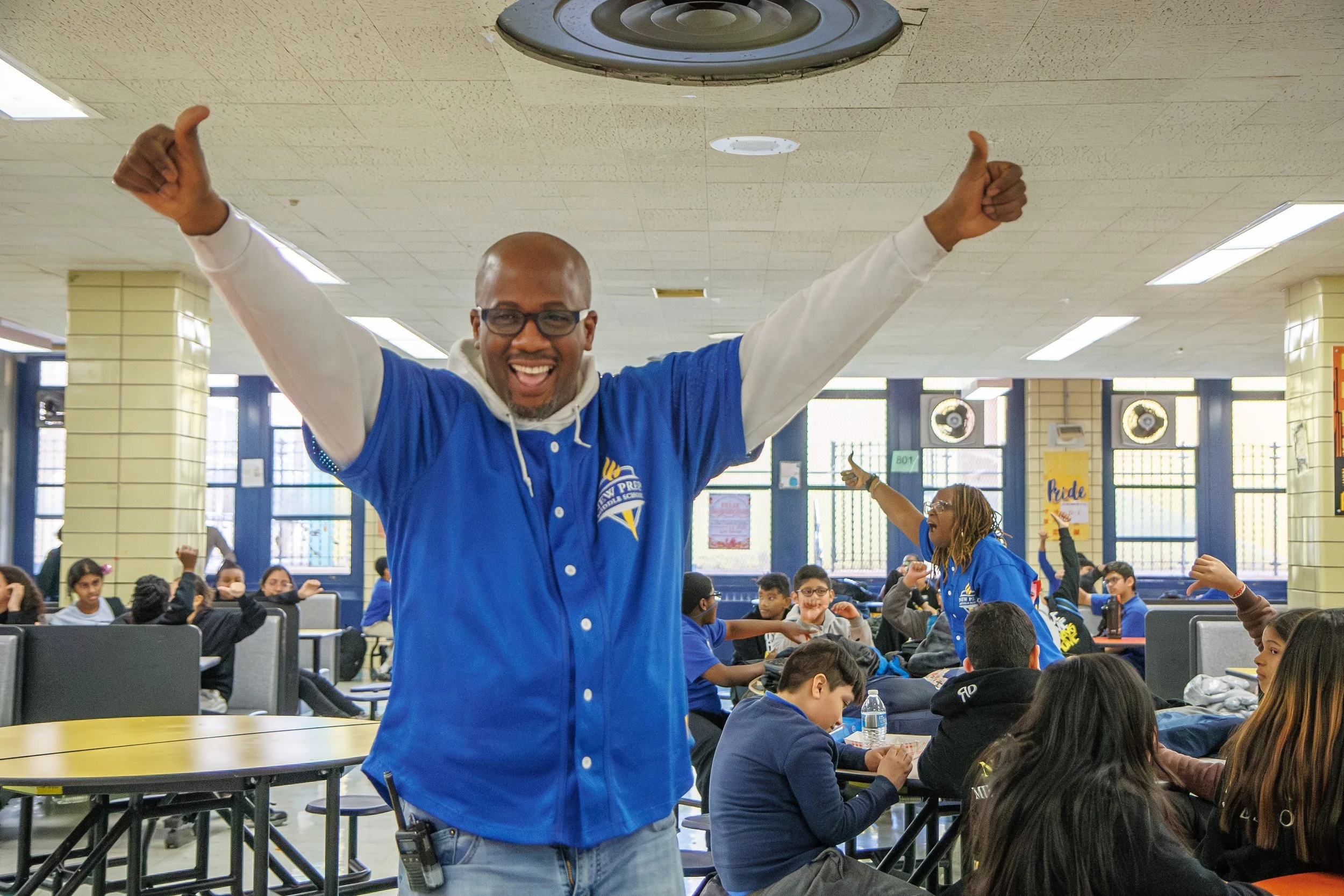 Smiling staff member in a blue shirt giving thumbs up in a busy cafeteria
