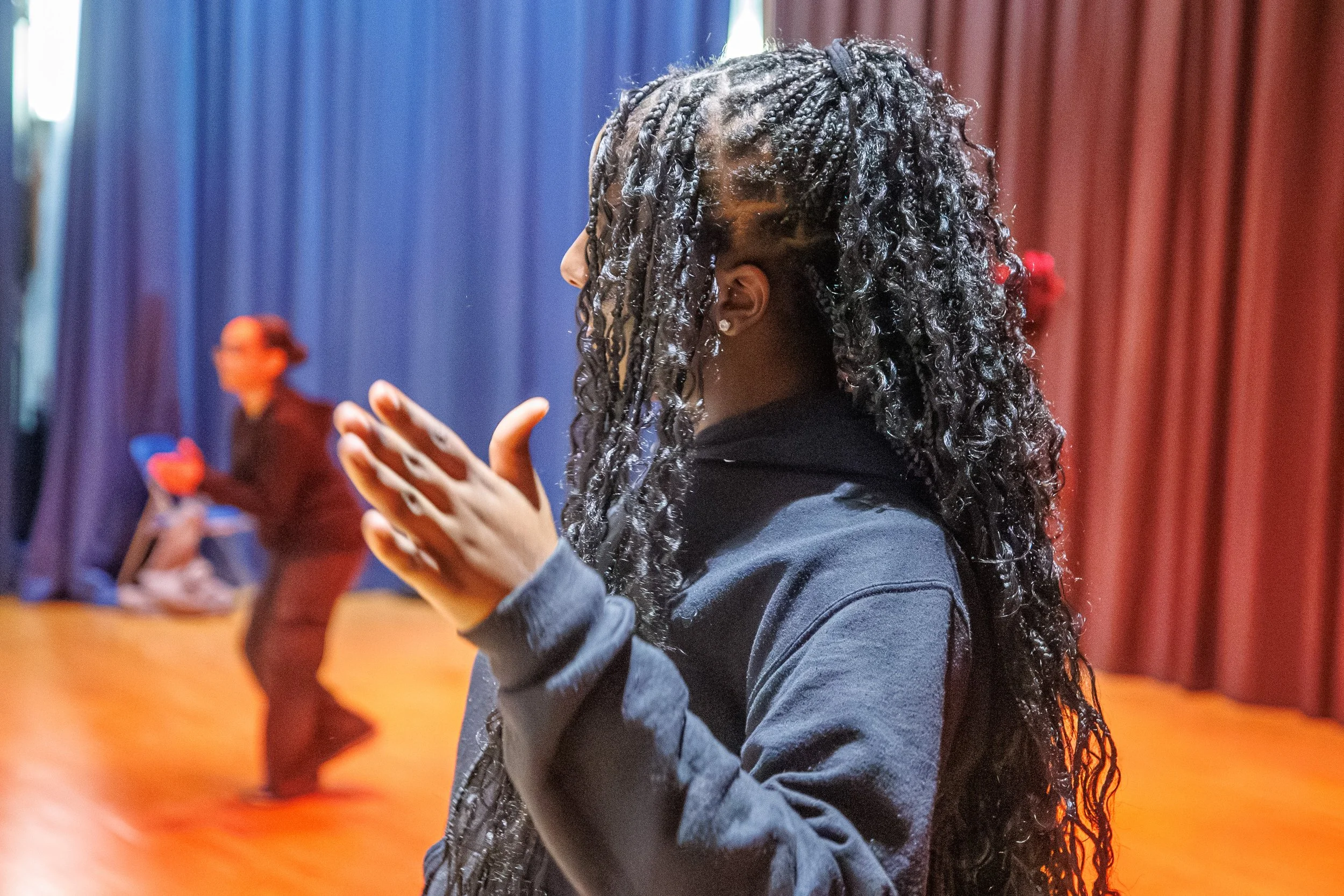 A person with long curly hair clapping in a gym or auditorium with blue and red curtains in the background.