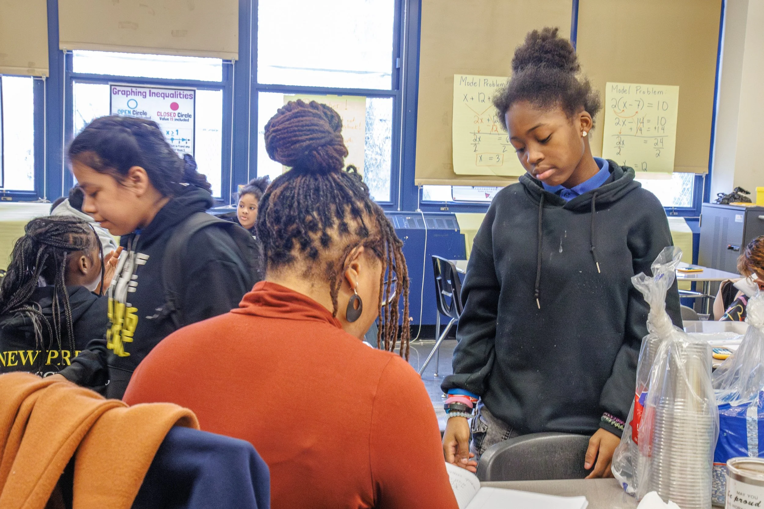 Students talking with a teacher at a desk in a classroom.