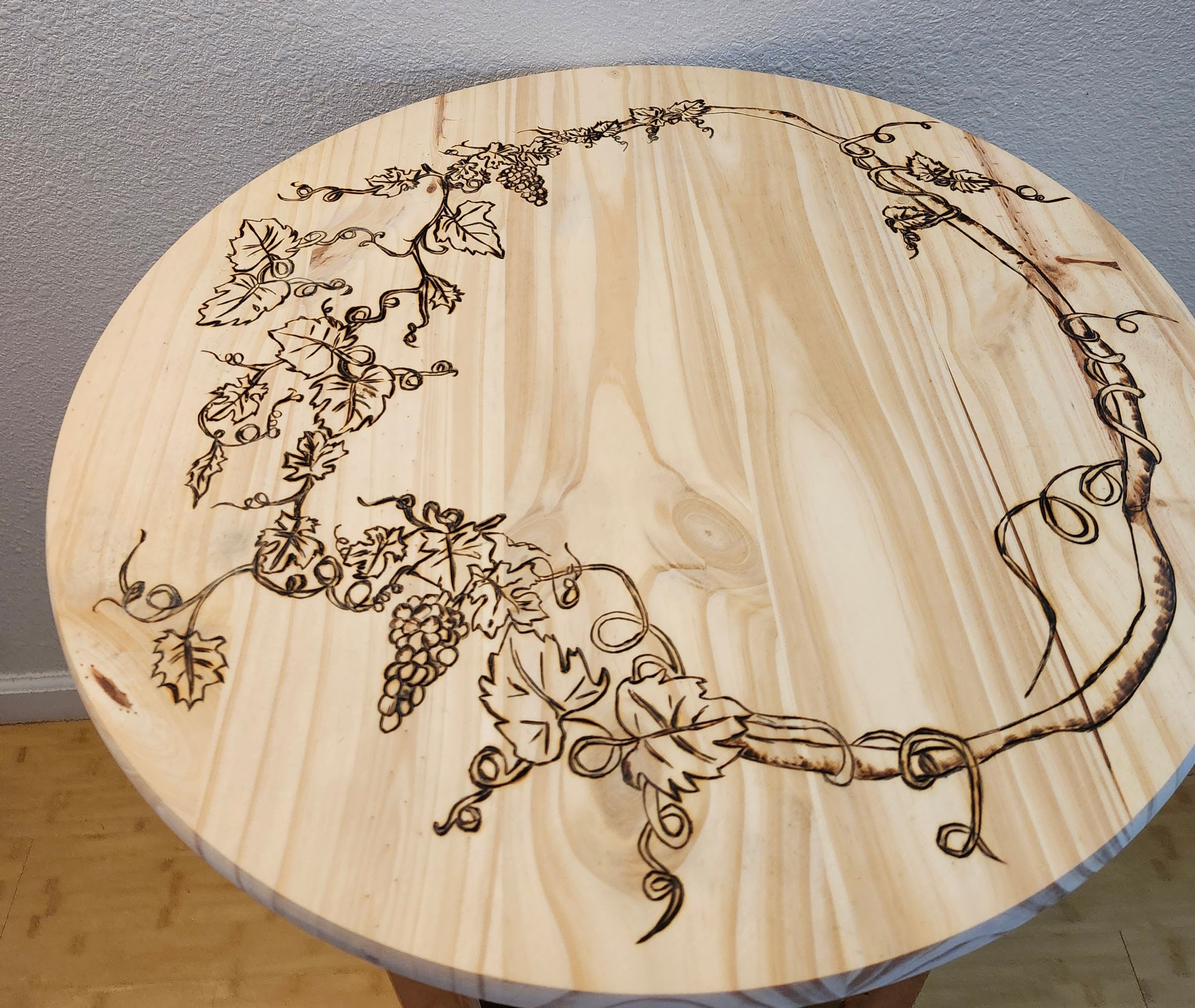 Round wooden table with a decorative vine and grape design burned into the surface.