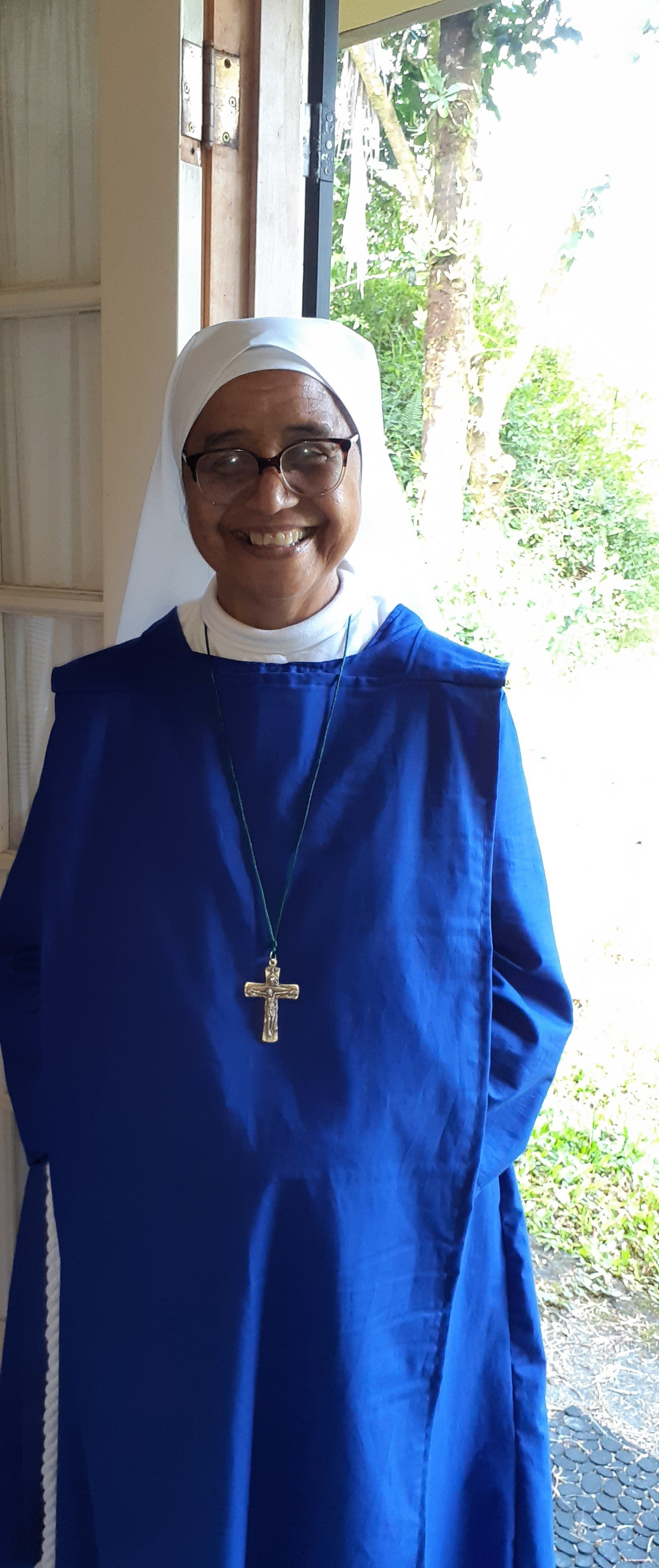 A smiling nun wearing glasses, a white habit, and a blue religious habit with a crucifix necklace, standing in front of a door with greenery outside.