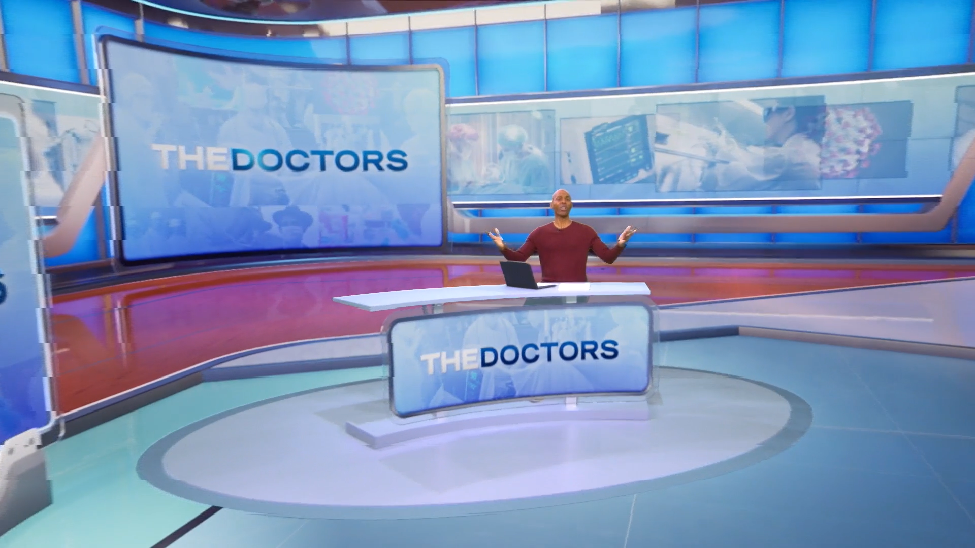 A television studio set with a large digital display screen showing 'The Doctors,' a person standing in front of it with arms raised, colorful background images, and a desk with the same logo.