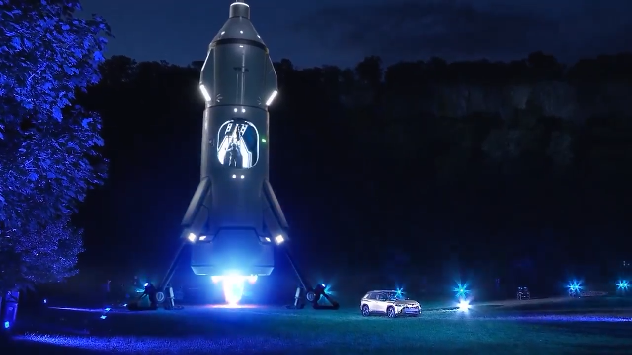 A large futuristic rocket standing upright outdoors at night, lit up with blue and white lights, with a small vehicle parked nearby and other lights in the background.