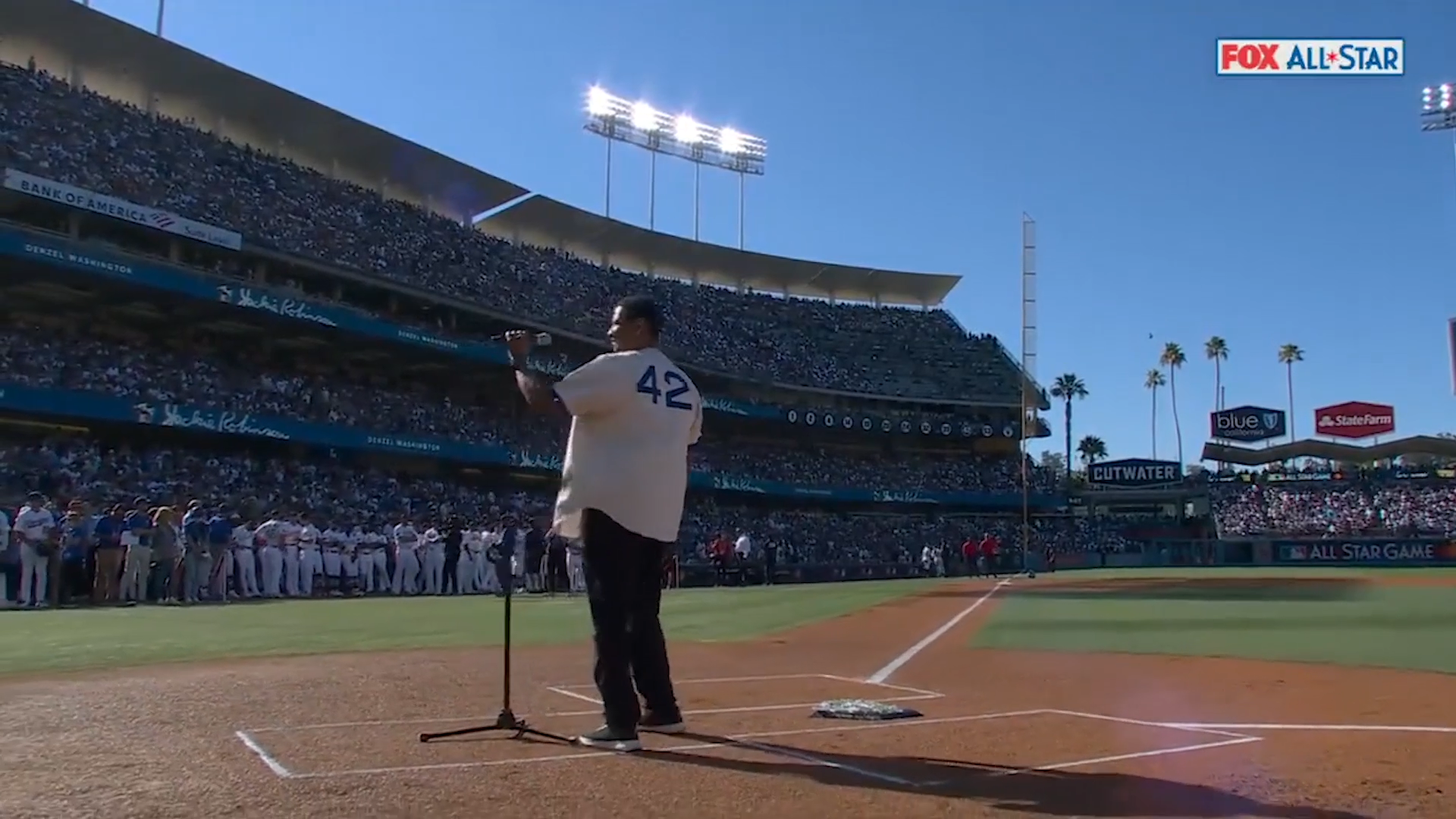 A baseball player stands at home plate during the All-Star game, holding a bat, with spectators in the stands and team members lined up along the first baseline. Bright sunlight shines in a clear blue sky.