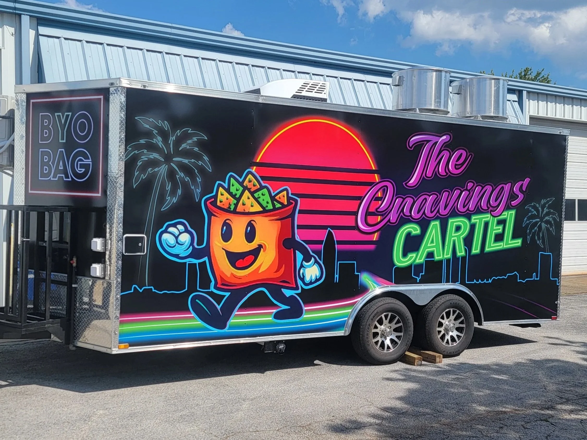 black concession trailer that has a bright colored wrap, a cartoon bag of chips open and walking in front of a sunset, text reads The Cravings Cartel