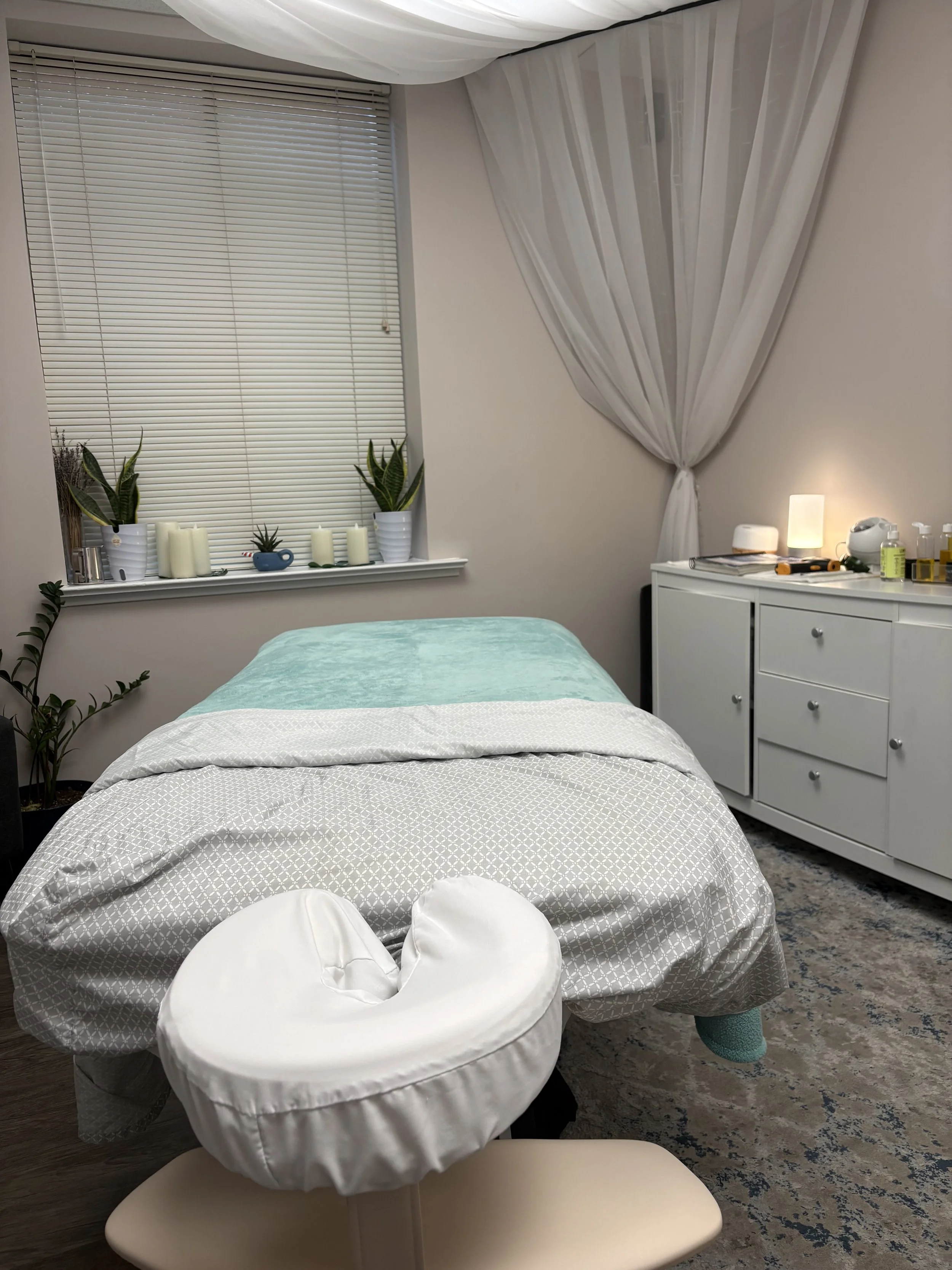 A massage or therapy room with a bed covered in a white and gray patterned sheet, a white circular pillow, and a beige massage chair. There are two potted plants, candles, and decorative items on a windowsill. The window has white blinds and sheer curtains. To the right, a white dresser holds a small lamp, a diffuser, and bottles of oils or lotion. The room has a peaceful, minimalist decor with soft lighting.