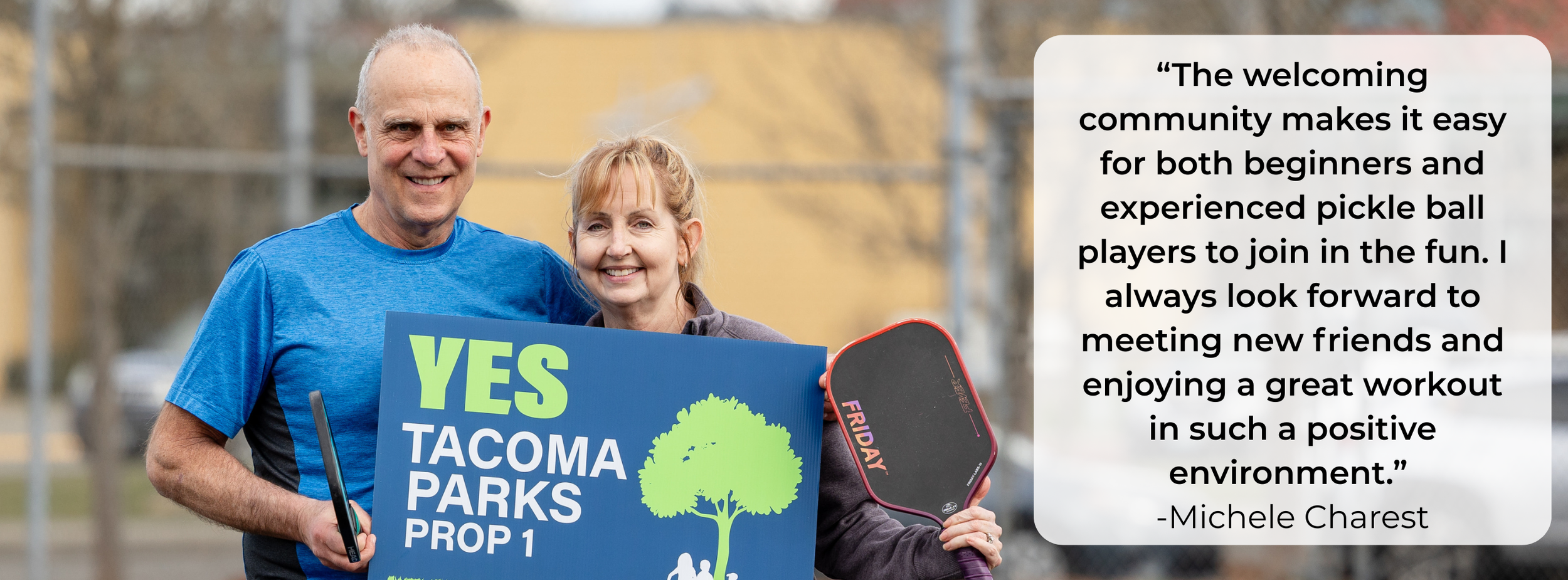 A smiling man and woman standing outdoors during the daytime, holding a sign promoting Tacoma parks and a pickleball paddle. The man is wearing a blue shirt, and the woman is wearing a gray jacket. They appear to be part of a community or recreational event.