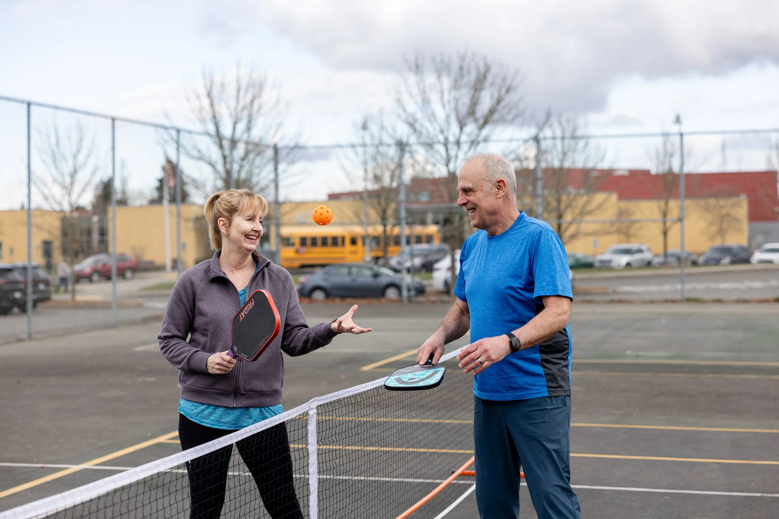 A woman and a man playing pickleball on an outdoor court. The woman is holding a paddle and smiling, while the man is holding a paddle, smiling, and seems to be hitting an orange ball.