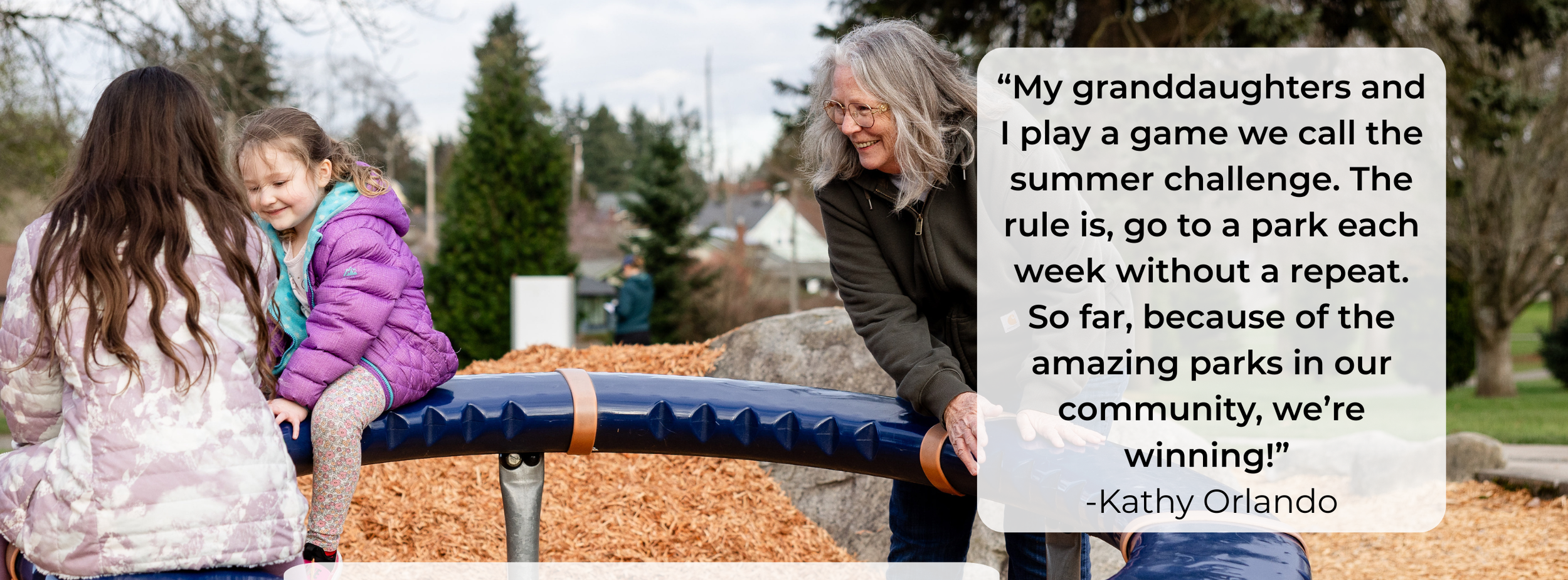 A woman with gray hair and glasses talking to two young girls at a park, with trees and a person in the background, while they play on a large, round outdoor game.