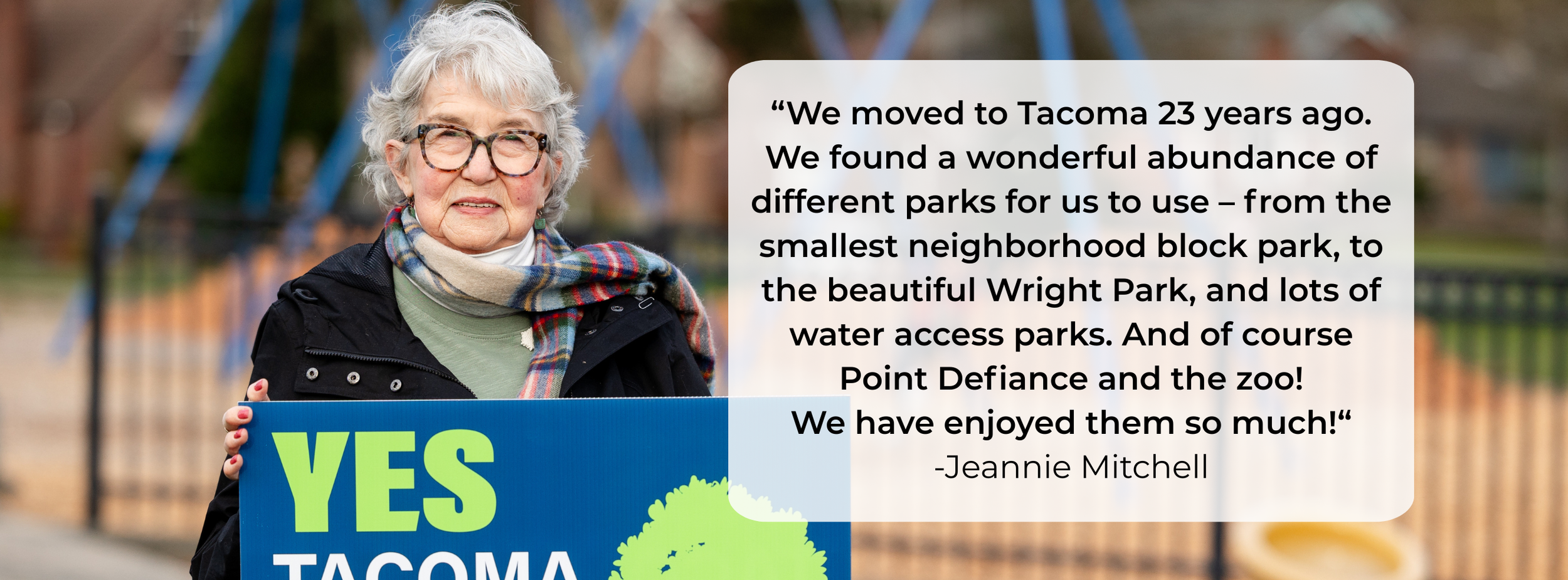 An elderly woman with glasses and gray hair holding a blue sign that reads "YES TACOMA". She is outdoors in a park with a playground in the background, wearing a dark jacket and a colorful scarf.