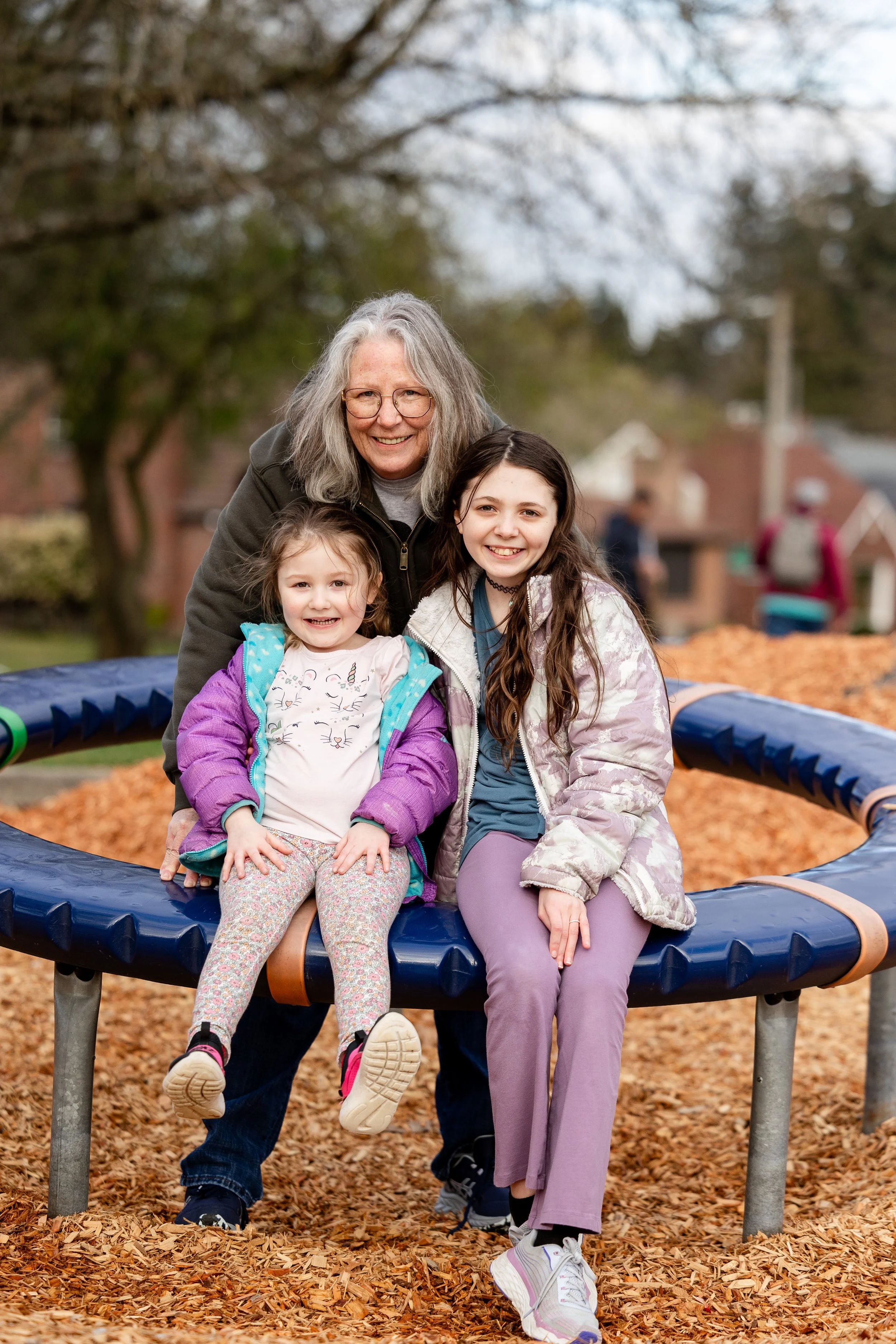 A woman and two young girls smiling and sitting on a large round playground structure in a park with trees and other children in the background.