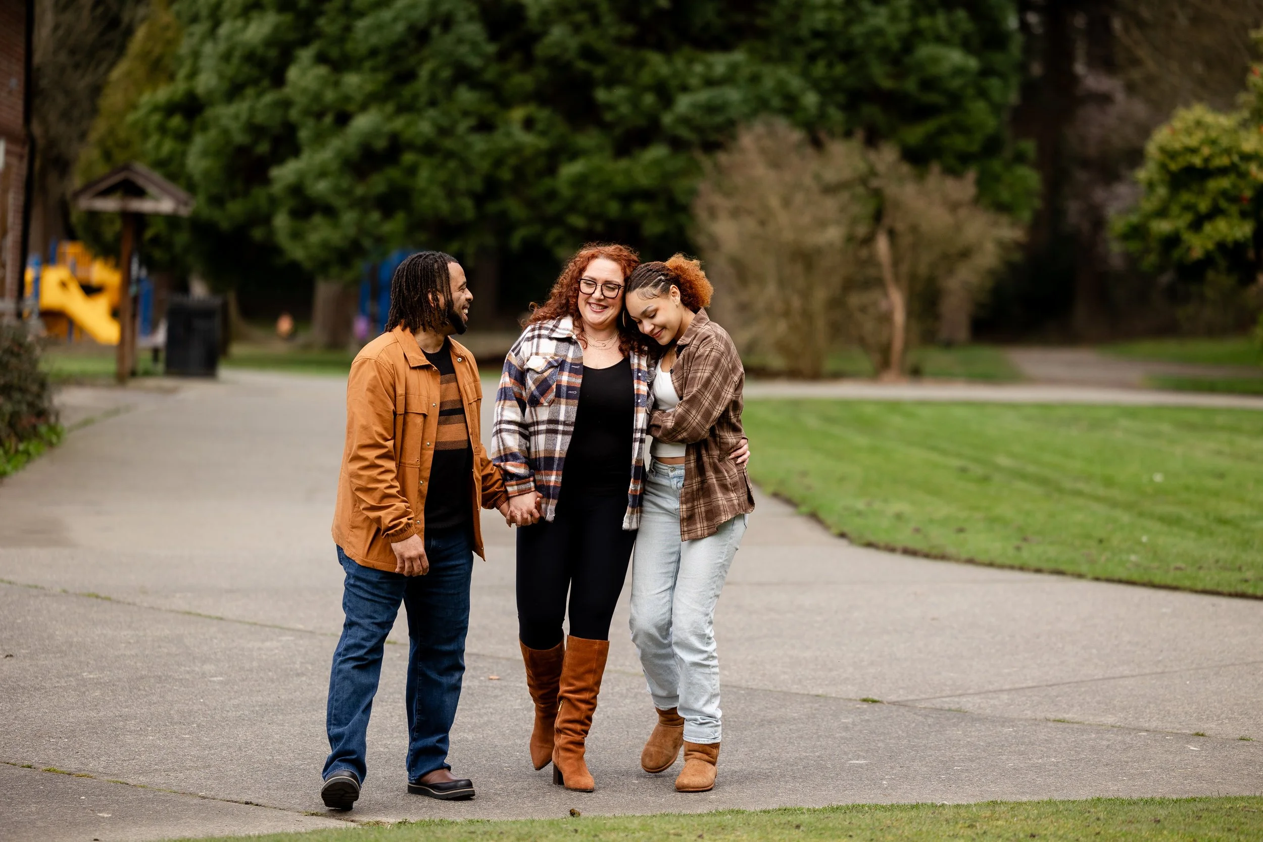 Three people walking together in a park, smiling and holding hands, with a playground and trees in the background.
