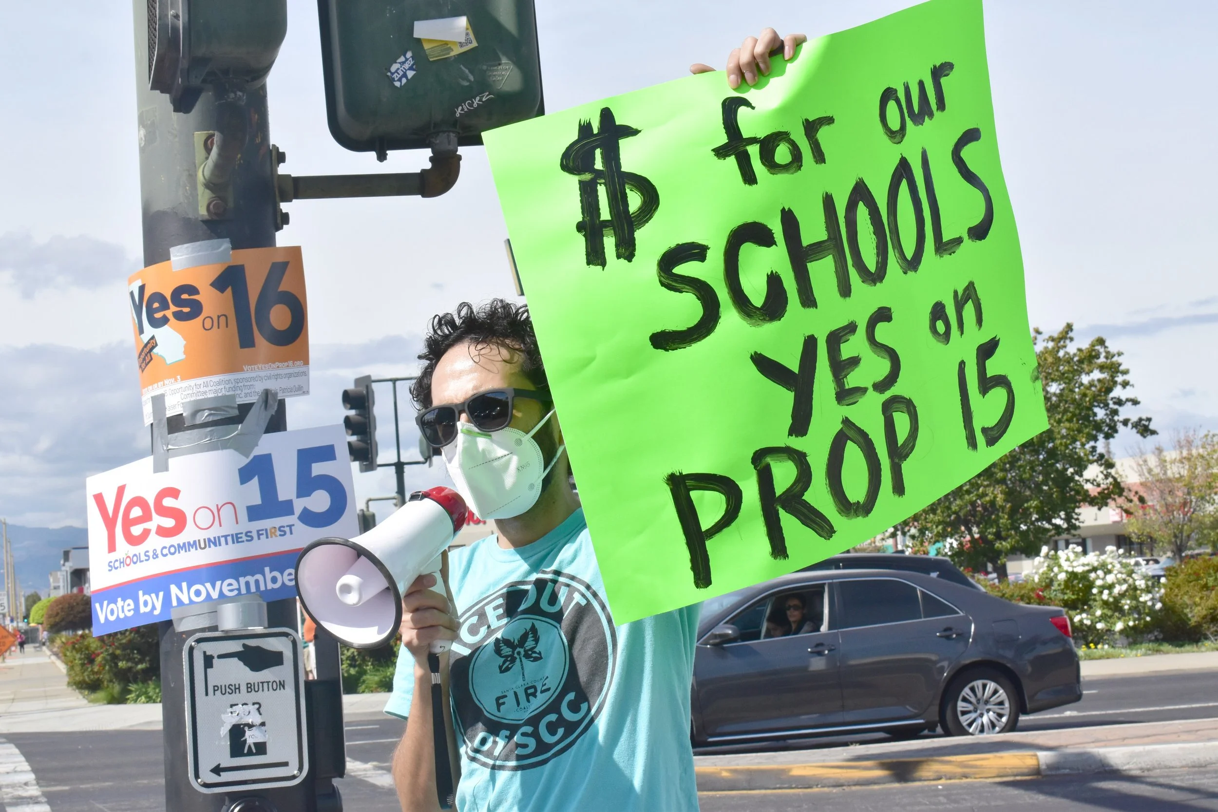 Person wearing a mask and sunglasses holding a green protest sign that reads '$$ for our schools' with a dollar sign symbol and other text. They are standing near a traffic light and other signs during a demonstration, with cars and trees visible in the background.