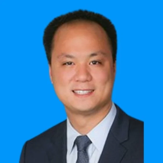 A professional headshot of a man with short black hair, wearing a dark suit and a tie, against a blue background.