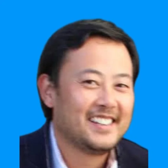 Close-up of a smiling man with black hair and a dark blazer against a blue background.