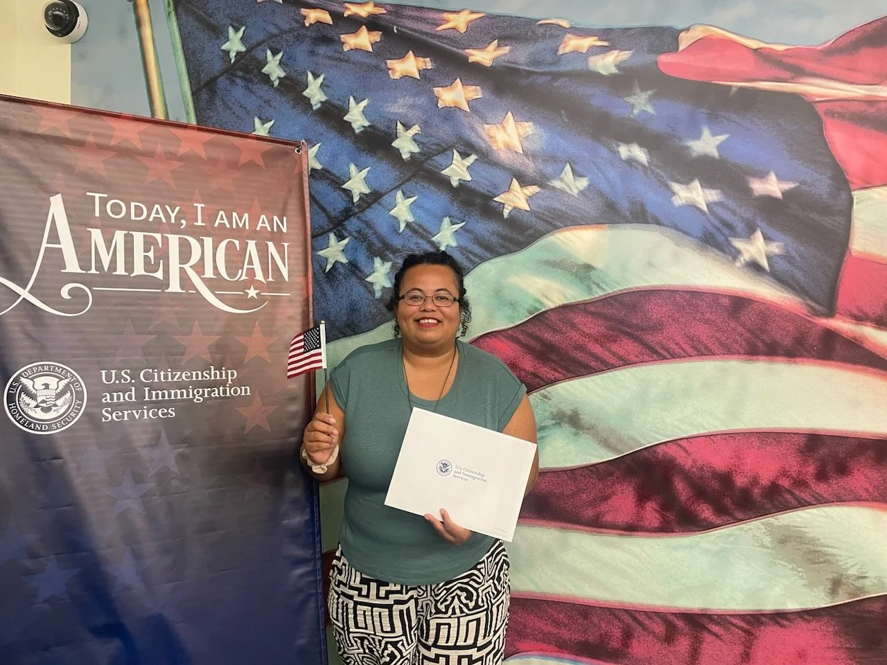 A woman smiling at a U.S. citizenship ceremony, holding an American flag and a certificate, standing in front of a large mural of the American flag with a patriotic banner that reads 'Today, I am an American.'