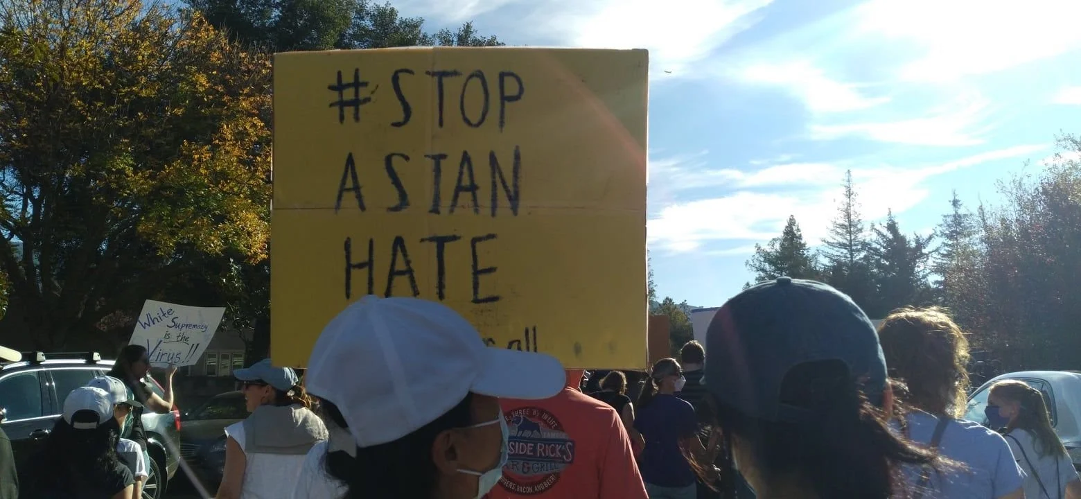 A group of people protesting outdoors, some wearing masks, with signs including a yellow sign in the foreground that reads '#STOP ASTAN HATE' and another sign in the background that reads 'White supremacy is the virus!!' under sunny skies.