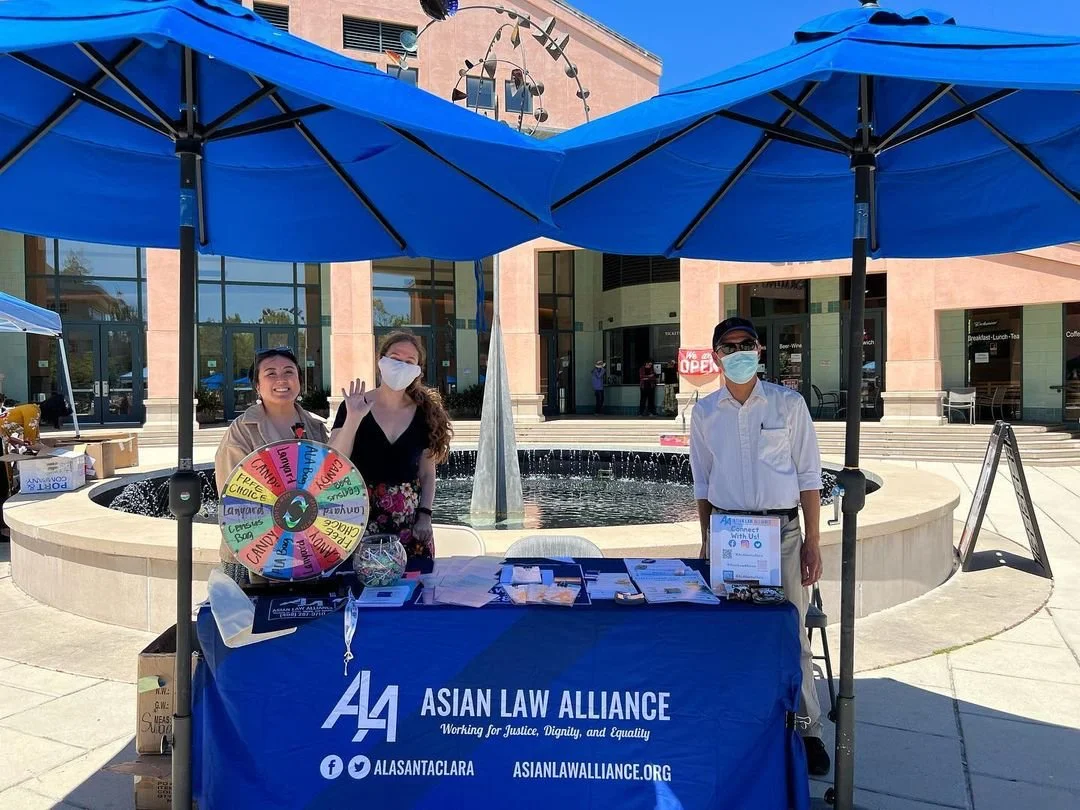 Three people standing behind a table with a blue tablecloth that says 'Asian Law Alliance' in front of a fountain and a building. The table has pamphlets, a prize wheel, and a sign-up sheet. Two people are wearing masks, and all are outdoors under blue umbrellas.