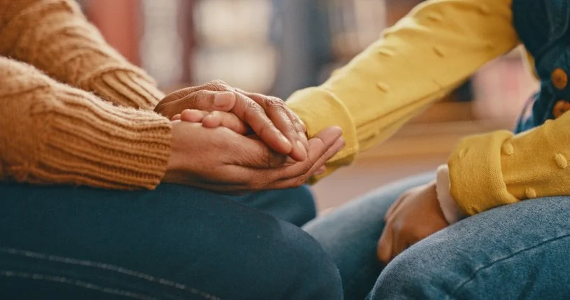 A person in a brown sweater holding another person's hand in a comforting gesture.
