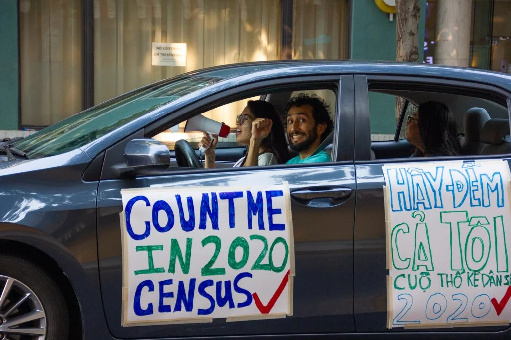 People sitting in a car participating in a census drive with signs saying 'Count me in 2020 census' and a message in Vietnamese encouraging people to participate in the census, on a city street.