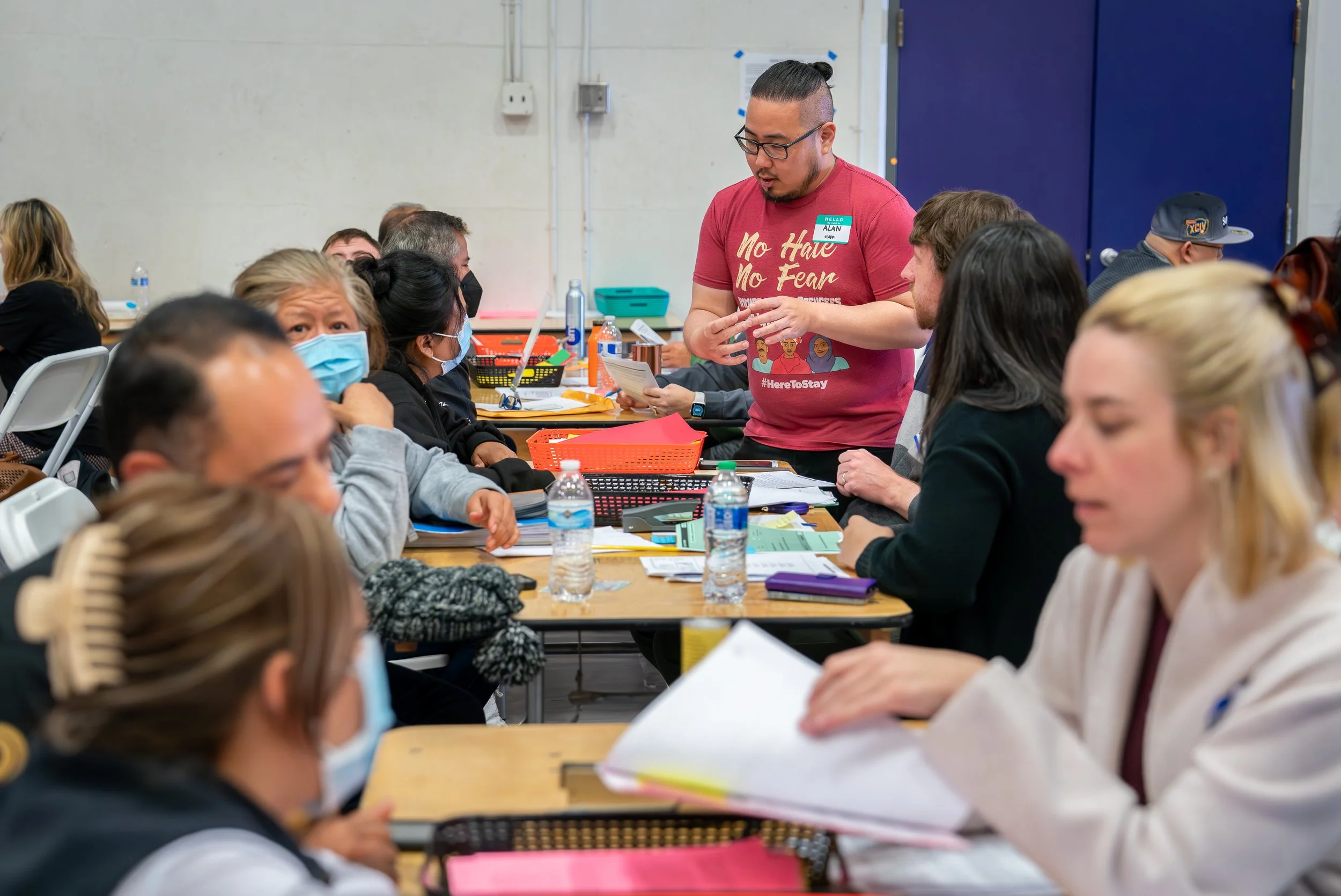 A diverse group of people sitting at tables, wearing face masks, engaged in a discussion or workshop. There is a man standing and speaking to the group, wearing a red shirt with text and a name tag.