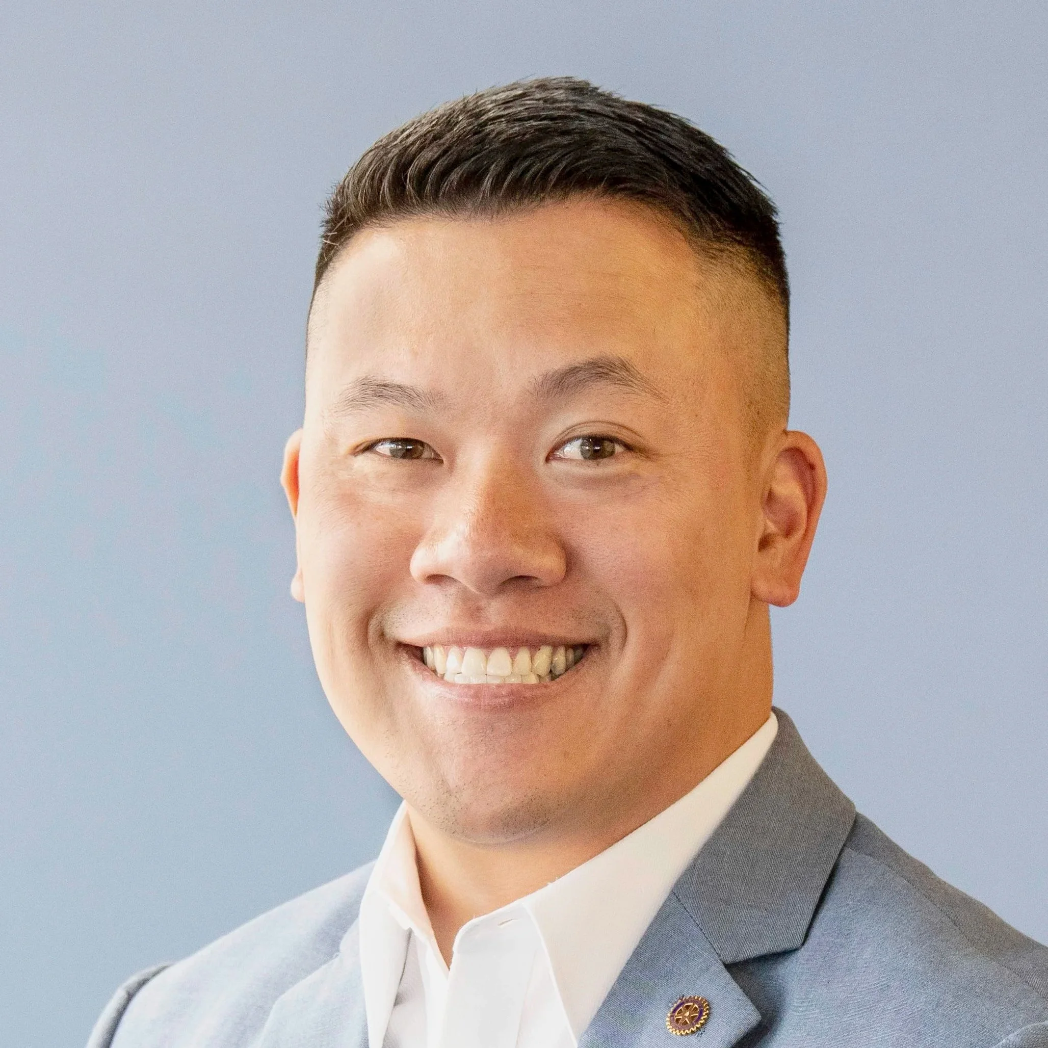 Close-up of a young man smiling, wearing a light gray blazer with a Rotary International pin and a white collared shirt, against a plain blue background.
