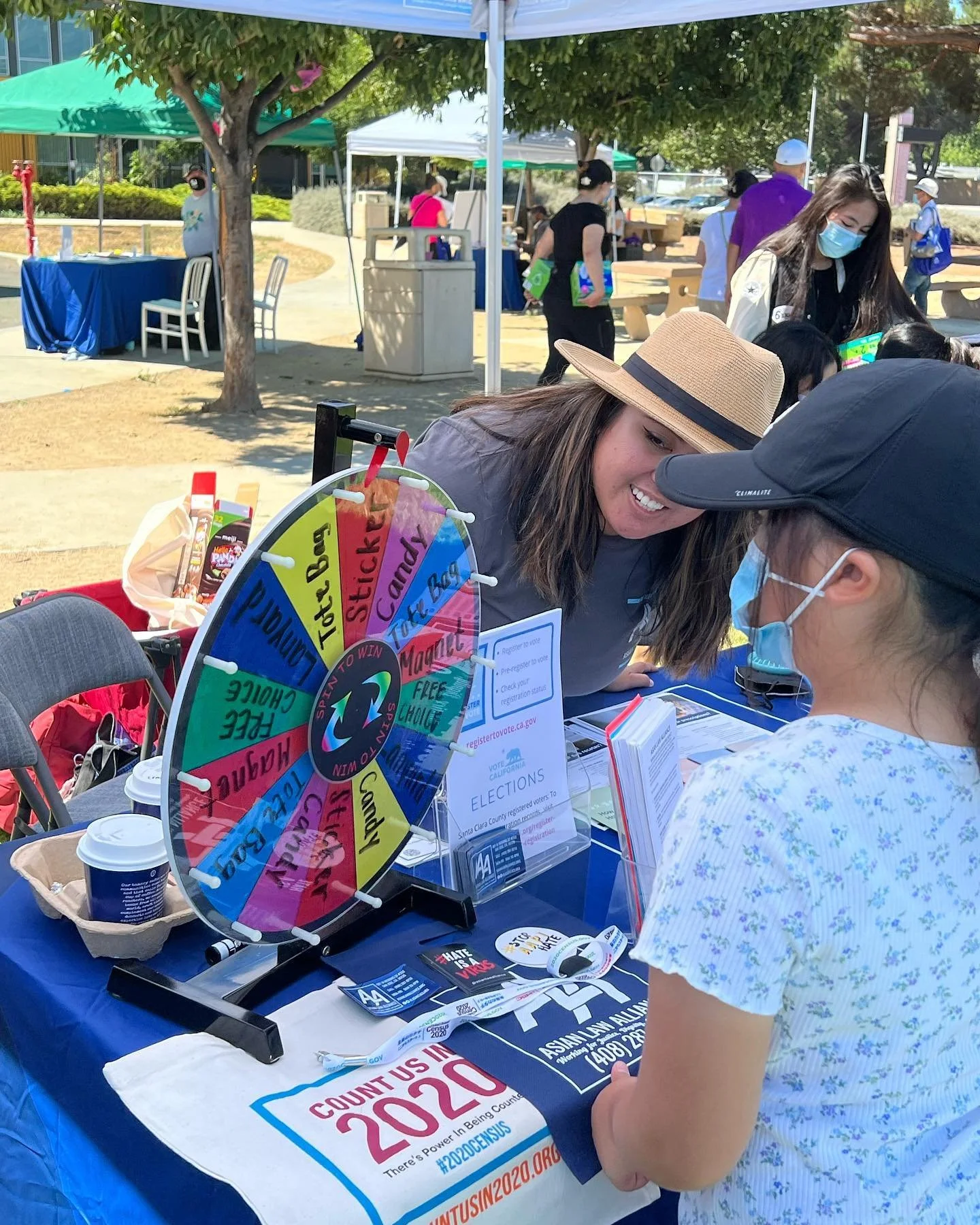Women at a table participating in a voting-related event outdoors, with a colorful spin wheel, informational flyers, and voting signs, under tents and surrounded by trees.
