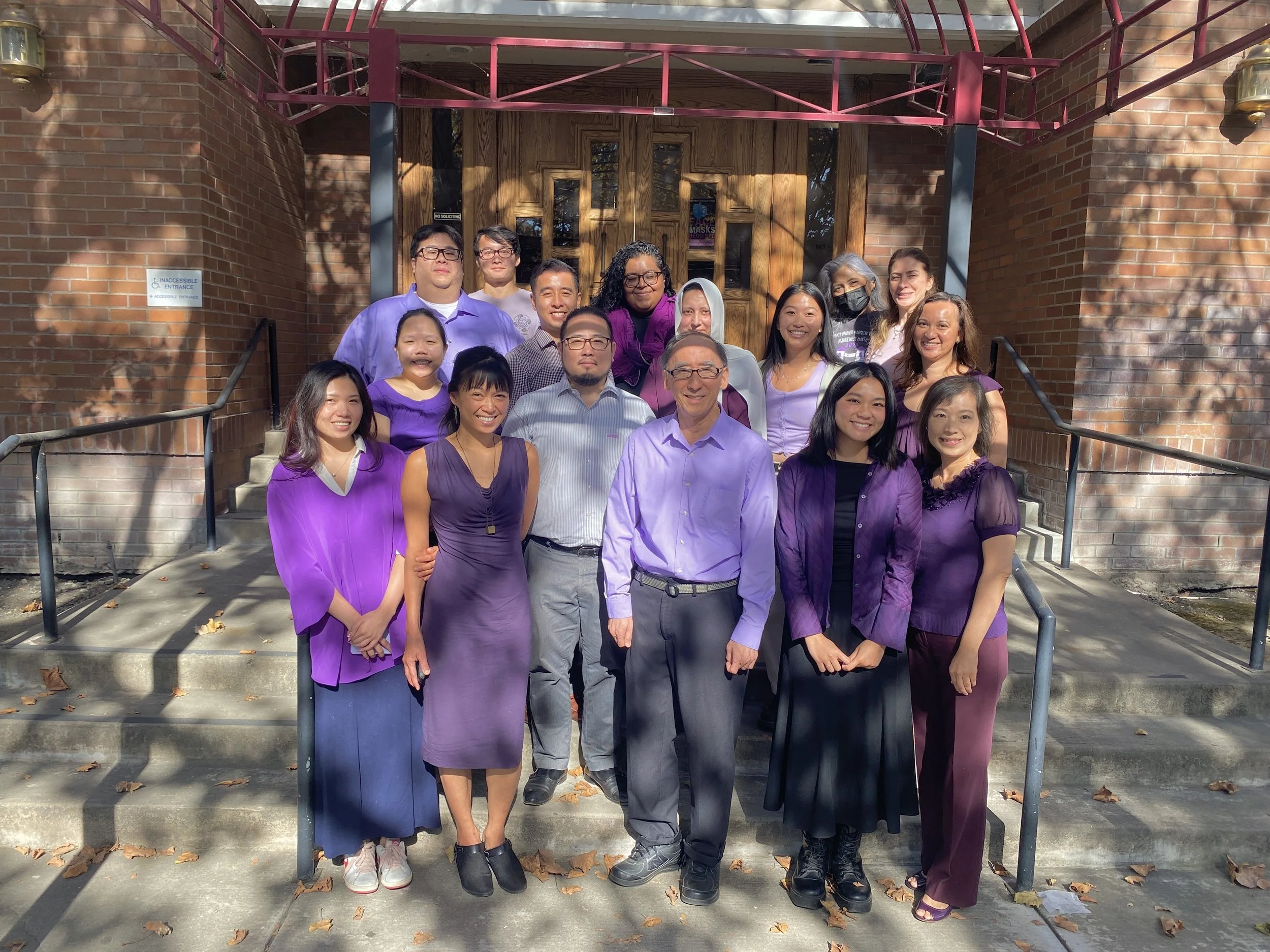 Group of diverse people standing on stairs outside a brick building, smiling and posing for a photo. Many are wearing purple clothing. There are trees casting shadows on the building and some fallen leaves on the ground.