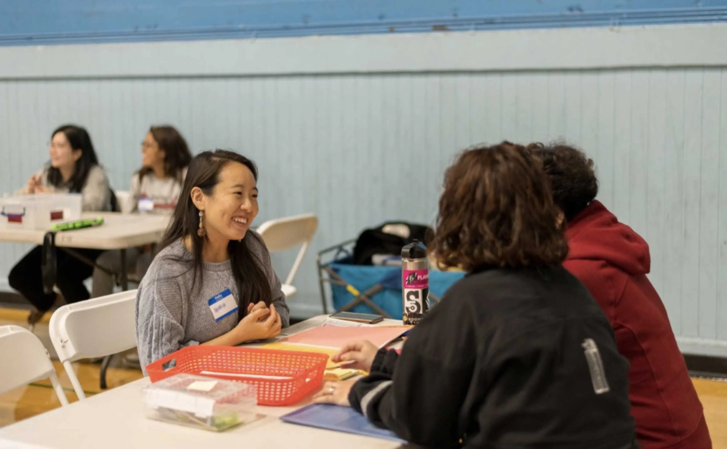 Group of women sitting at tables in a room, engaging in conversation and smiling, with Iced Coffee, stationery, and books on the table.