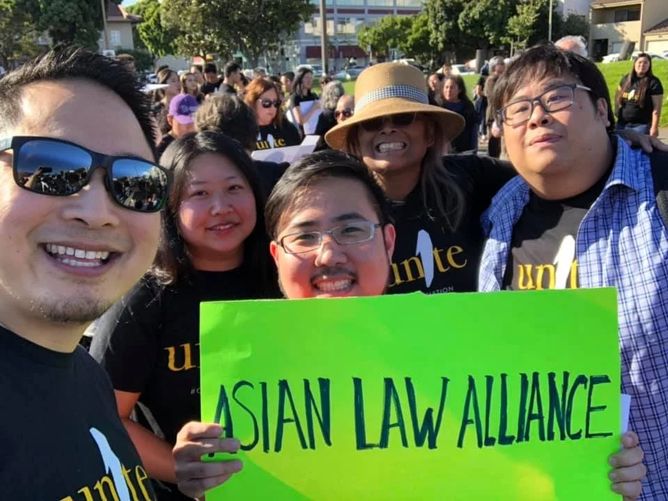 Group of diverse people at a rally holding a bright green sign that reads 'ASIAN LAW ALLIANCE', with others holding signs in the background.