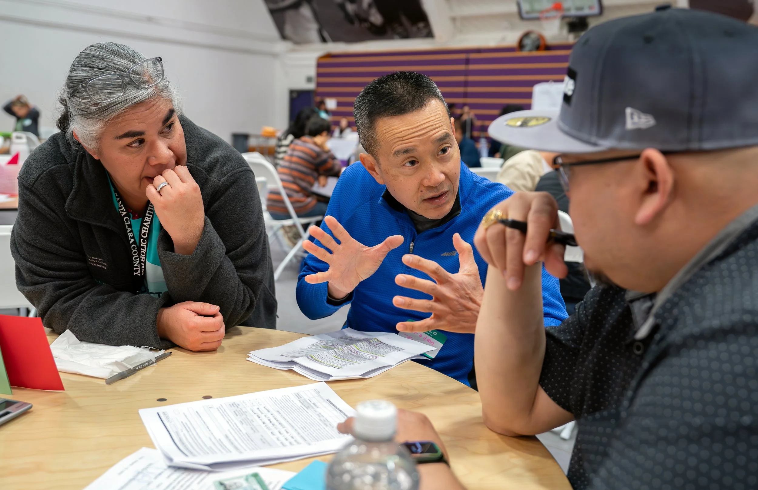 Three people engaged in conversation at a table filled with papers and documents, with a busy indoor setting in the background.