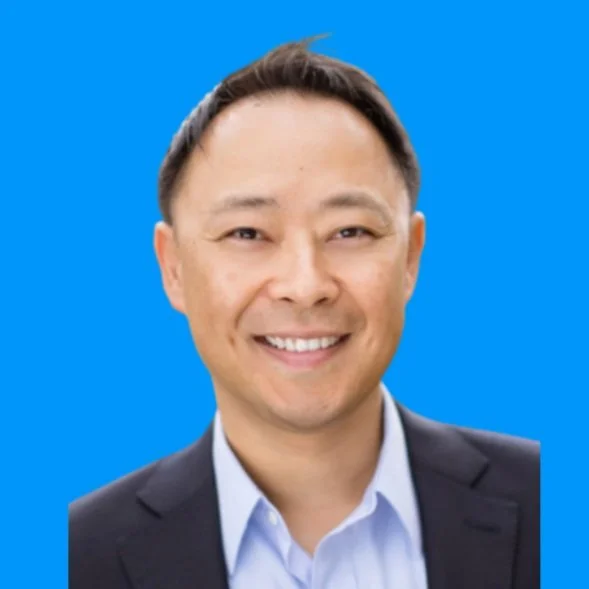 Professional headshot of a man smiling, wearing a suit and a light blue shirt, against a solid blue background.