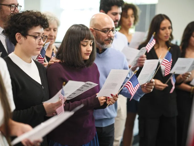 People of different ages and ethnicities participating in a patriotic ceremony, holding small American flags and reading from papers indoors.