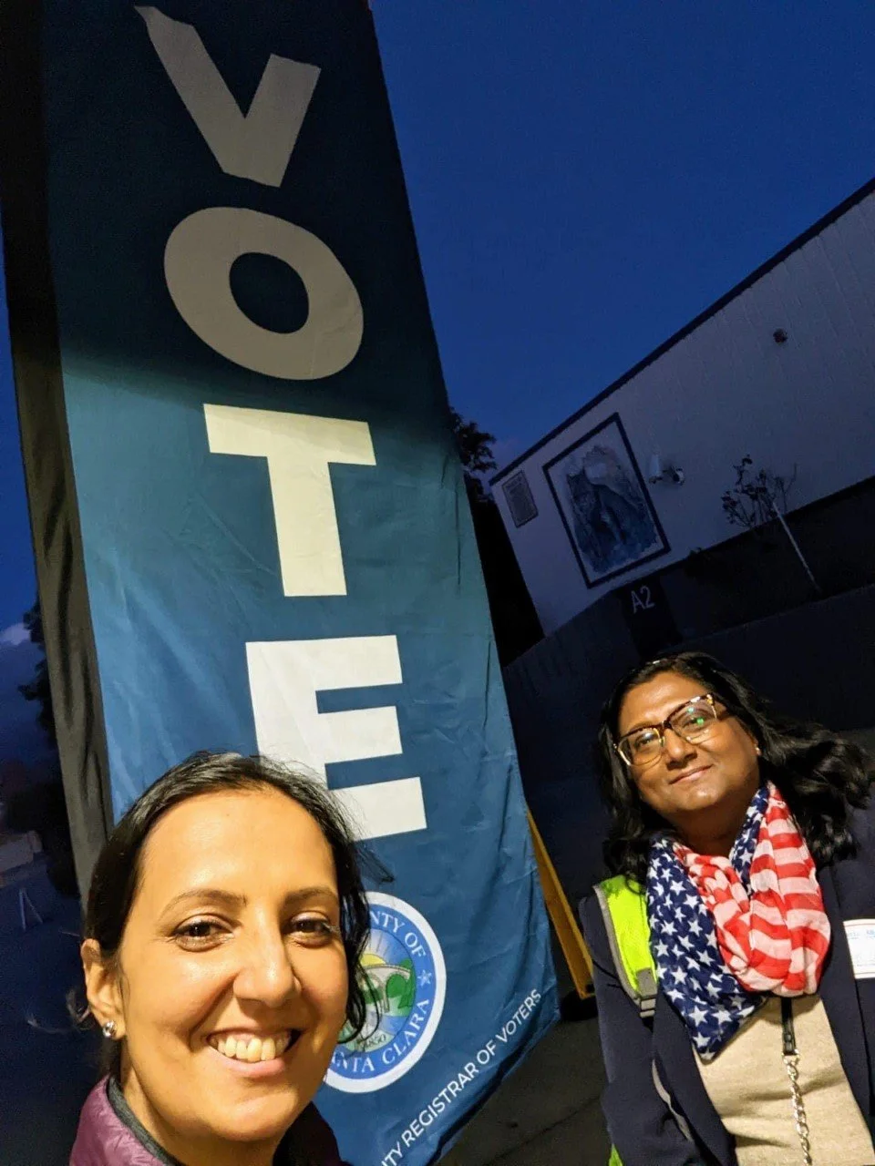 Two women taking a selfie near a large blue sign with the word 'VOTE' and a city seal, outdoors in the evening.