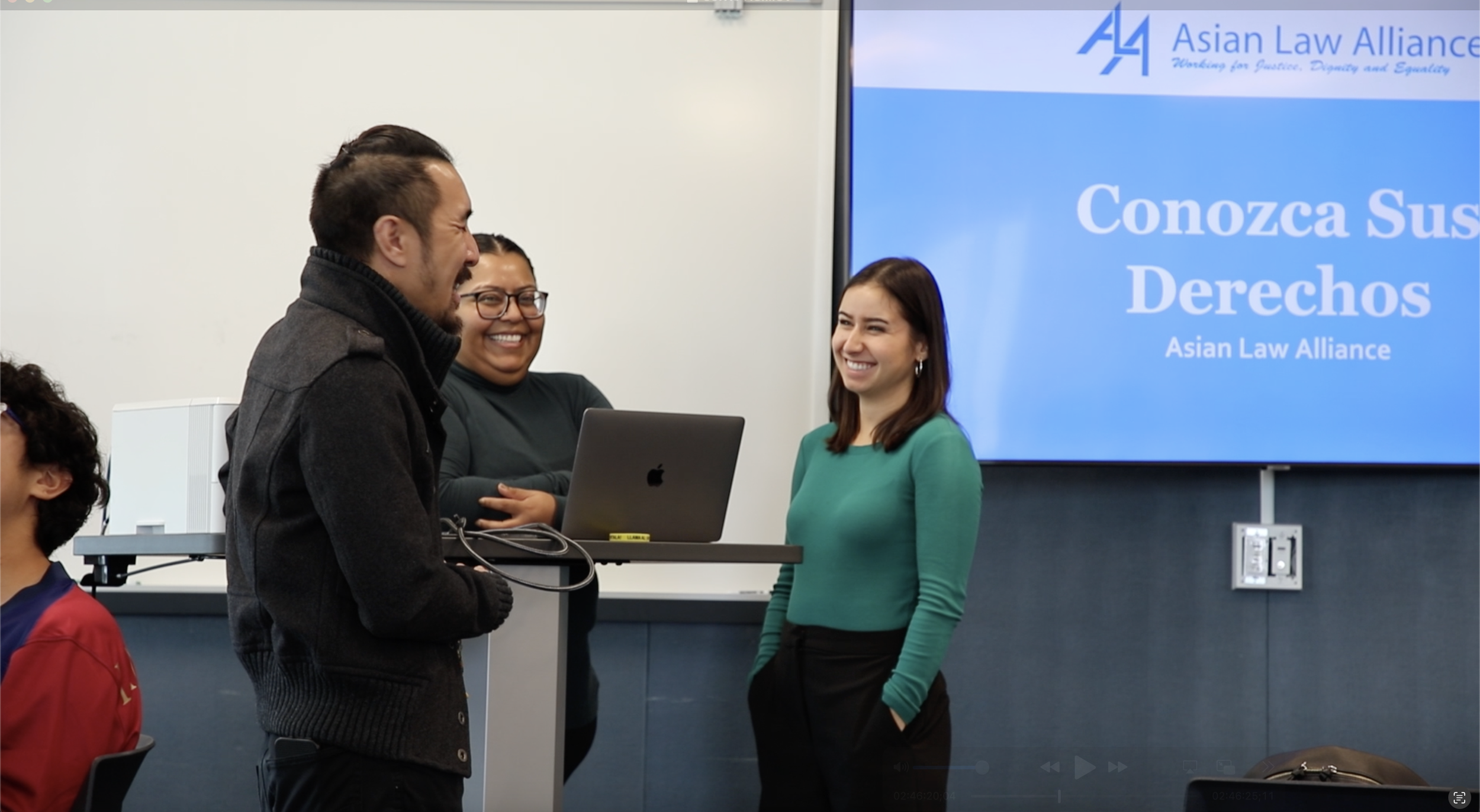 Three people are smiling and talking in a classroom or conference room. There is a large screen in the background displaying the Asian Law Alliance logo and text in Spanish about rights. One person is standing in front of a table with a laptop, and two others are standing nearby, one wearing glasses and another in a teal shirt.