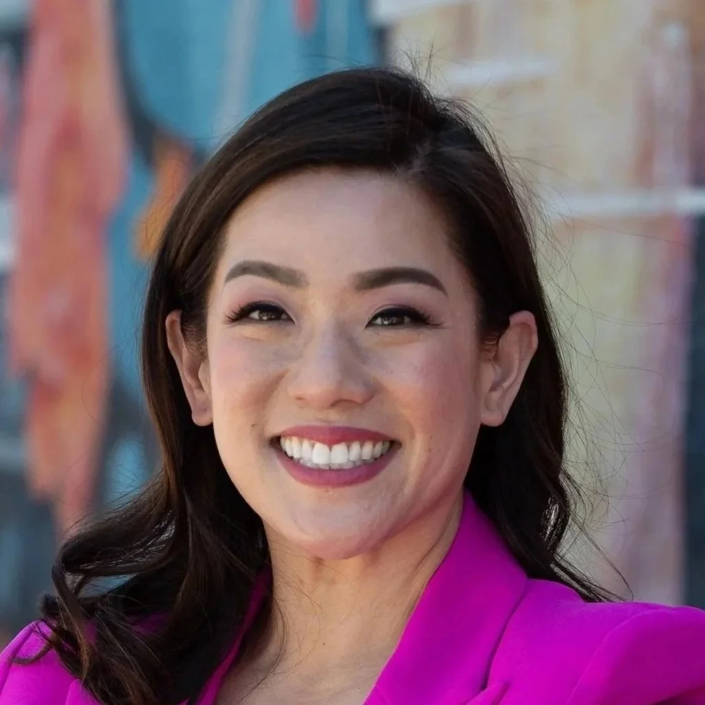 A woman with dark hair wearing a pink jacket, smiling at the camera, with colorful graffiti in the background.