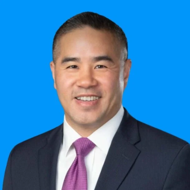 Professional headshot of an Asian man in a dark suit, white shirt, and purple tie, smiling against a blue background.