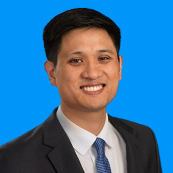 A professional headshot of a man with short black hair, wearing a dark suit and a tie, against a blue background.